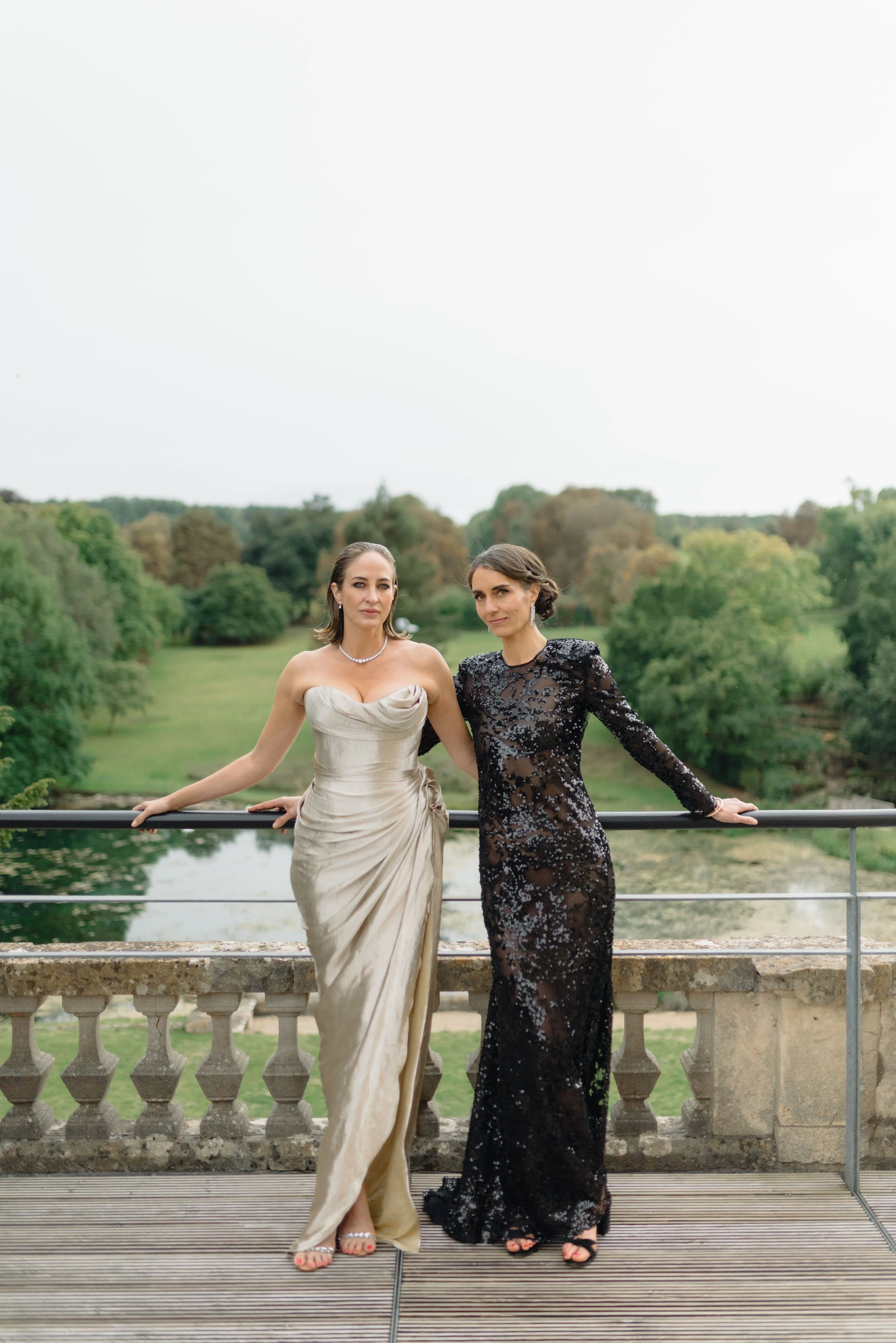 Two women in elegant evening gowns standing on a balcony with a scenic landscape of trees and a pond behind them.