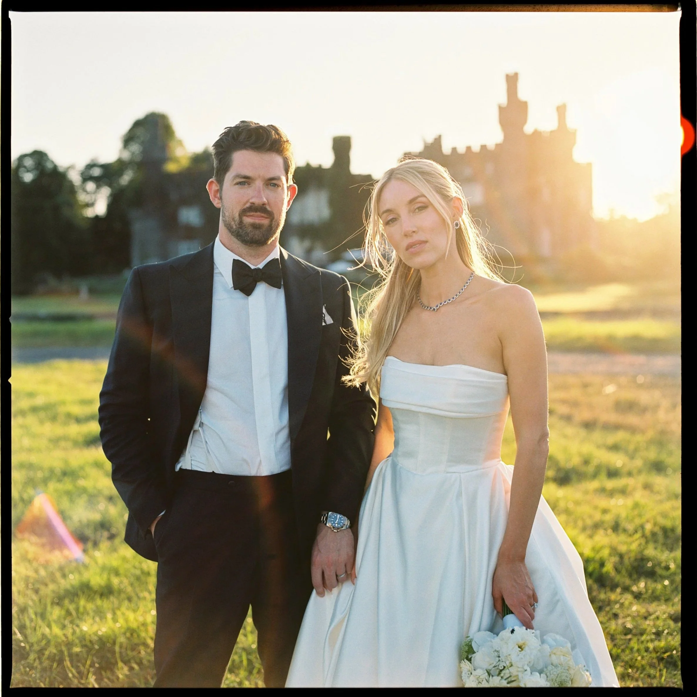 A groom in a tuxedo and a bride in a wedding dress standing outdoors on a sunny day, with Irish wedding venue Luttrellstown Castle in the background.