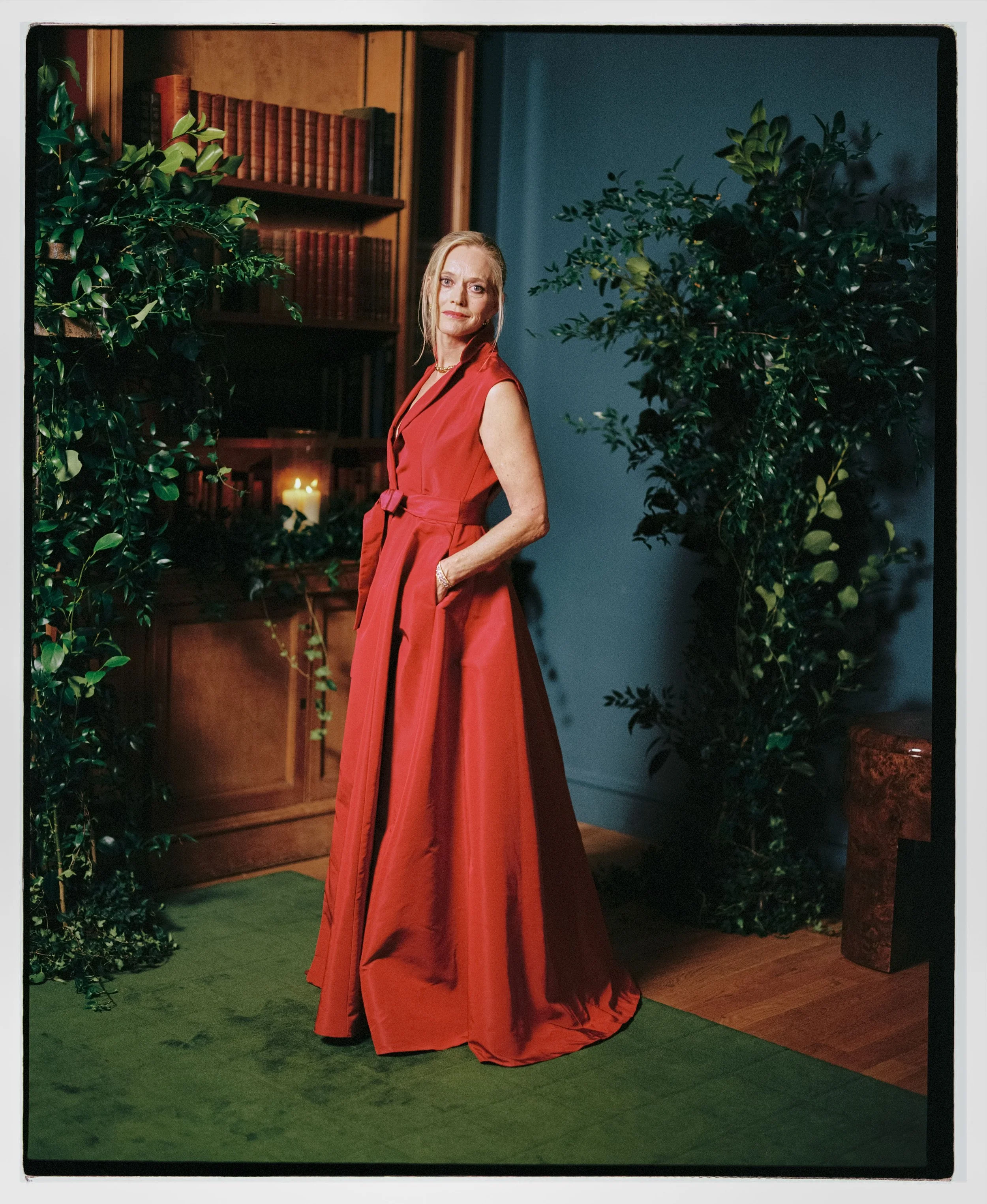 A woman in a long red dress standing indoors next to houseplants, a bookshelf, and a candlelit lantern.