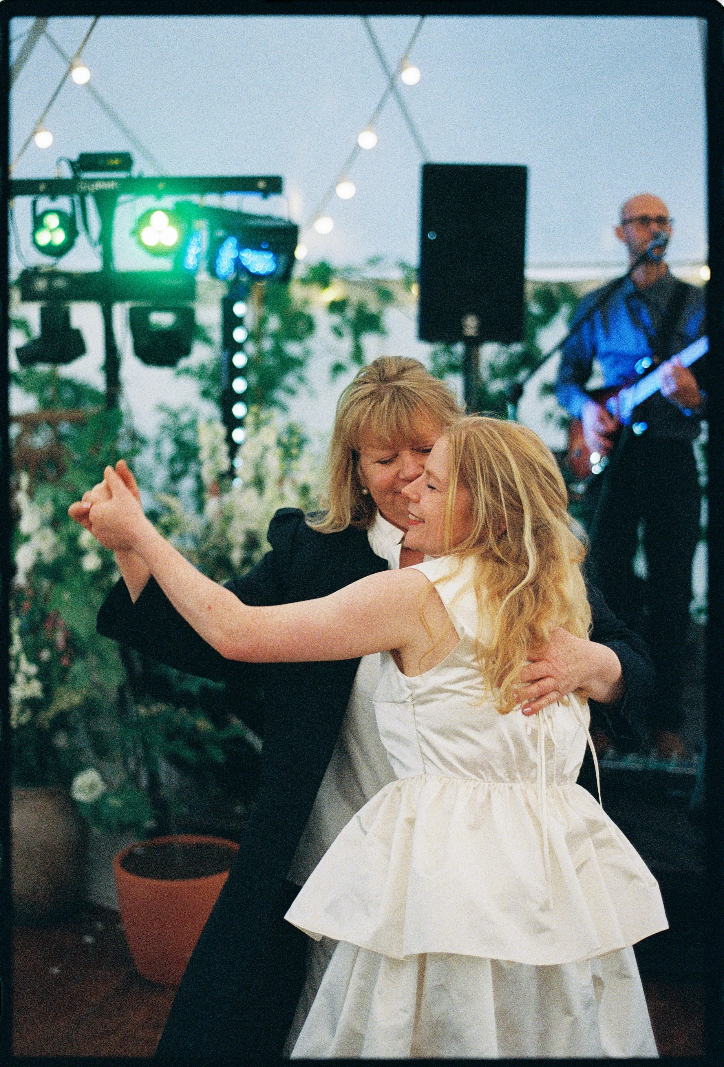 A woman and a girl are dancing closely together at a celebration, surrounded by string lights and a band playing music in the background.
