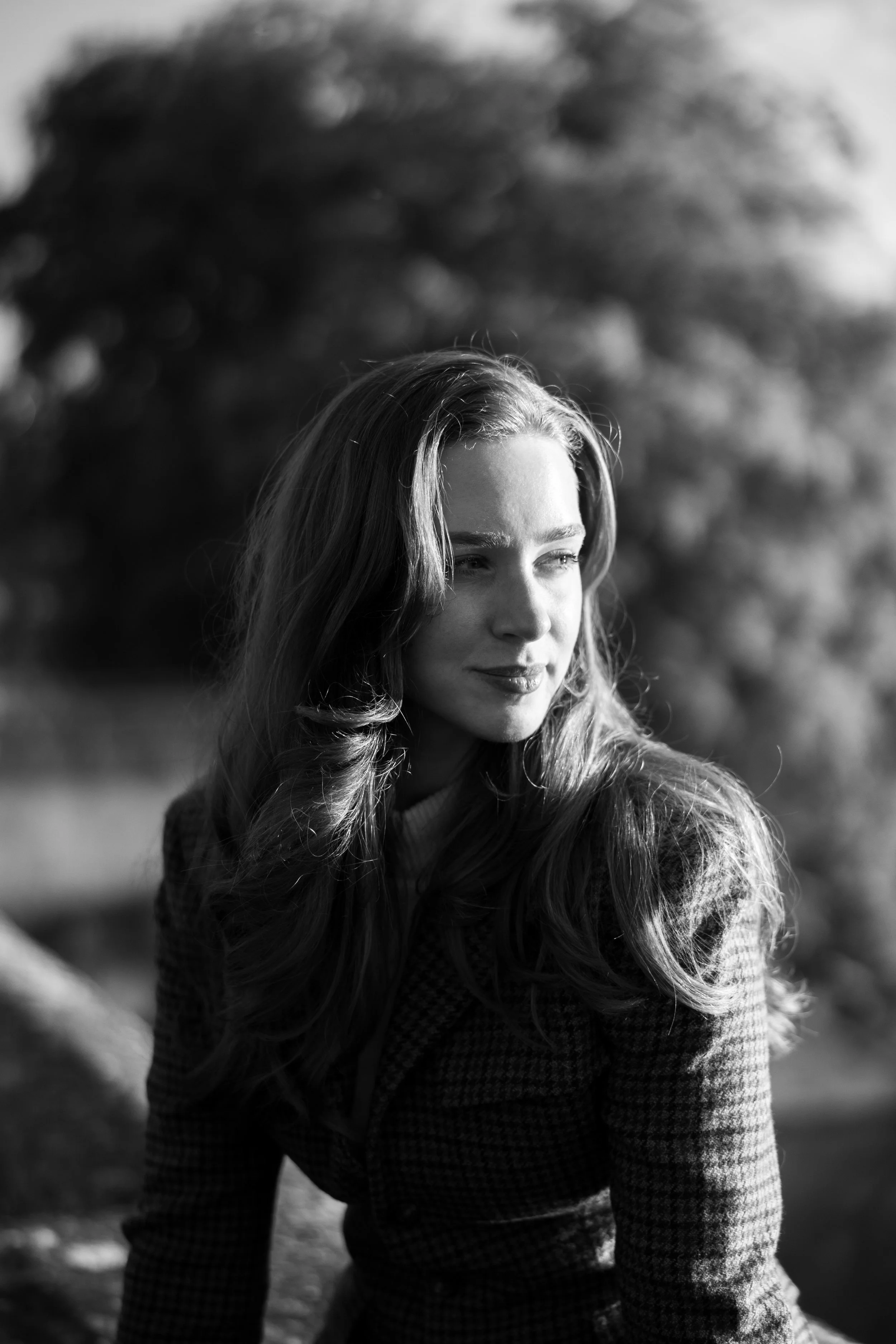 Black and white portrait of a woman with long wavy hair, looking off to the side with sunlight on her face and a blurred natural background.