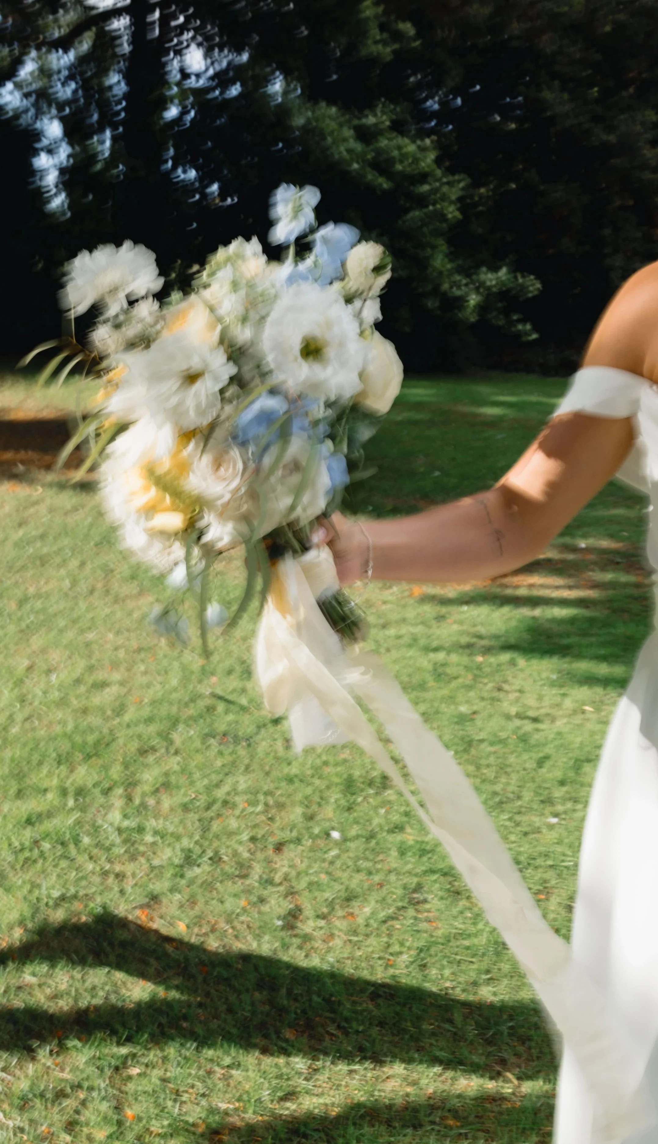 A bride holding a bouquet of white and cream-colored flowers with a ribbon, outdoors on grass with trees in the background.