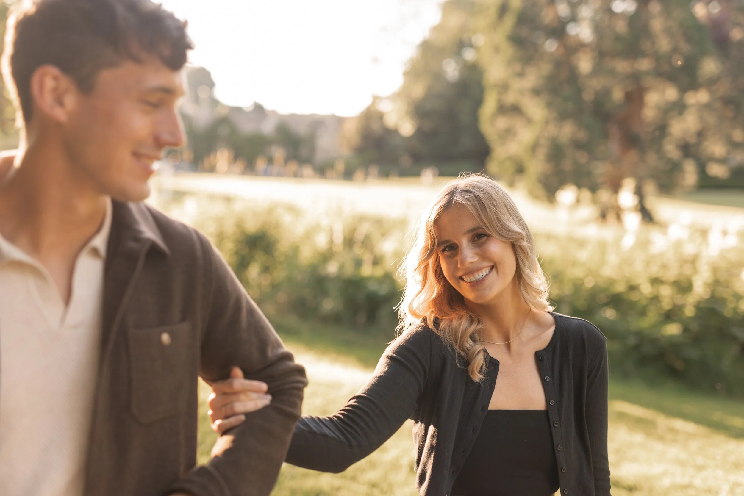 A woman smiling and holding hands with a man outdoors in a park during sunset.