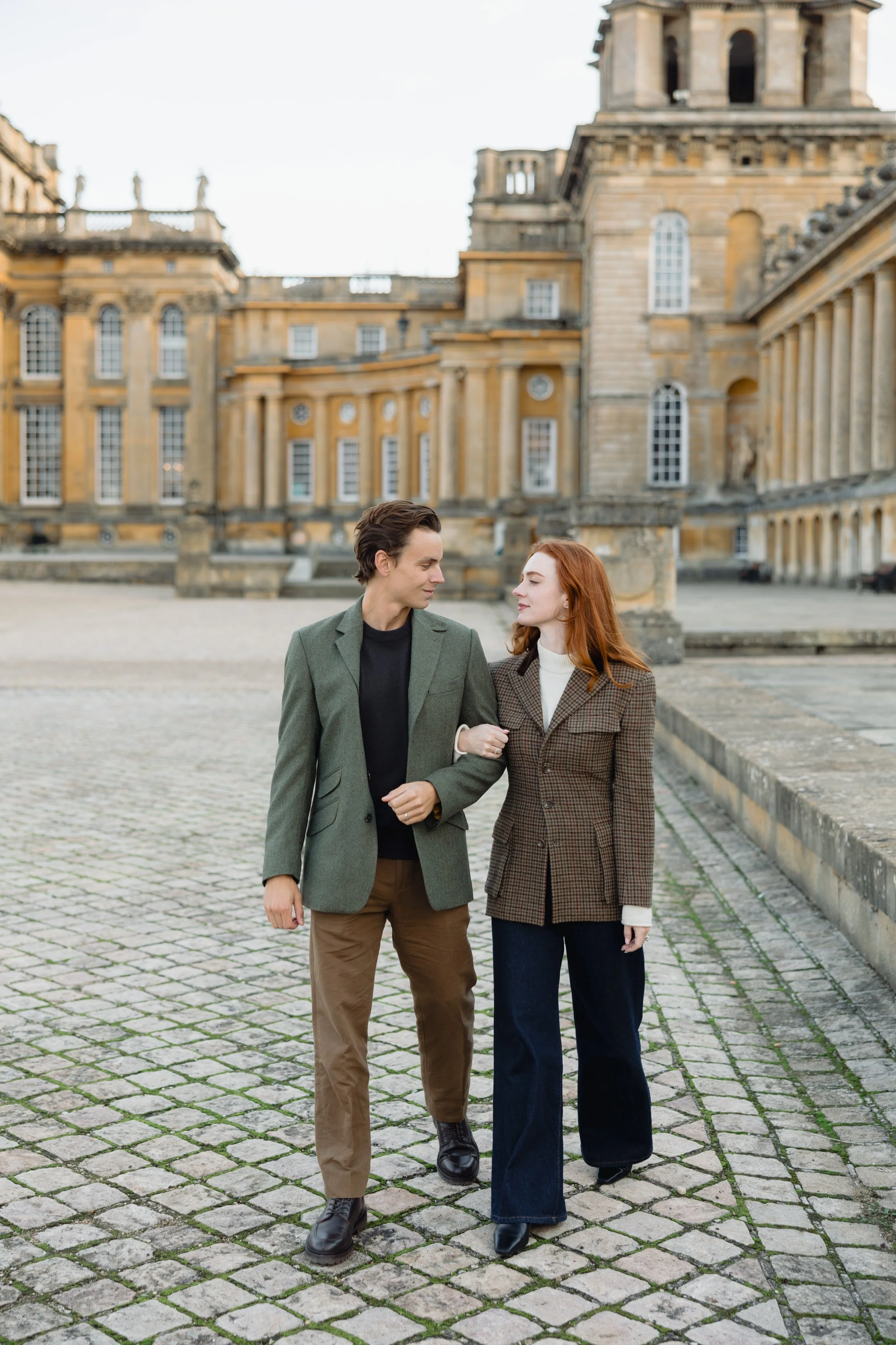 A young man and woman walking arm in arm on a cobblestone plaza in front of a historic building with multiple windows and columns, dressed in smart casual attire.