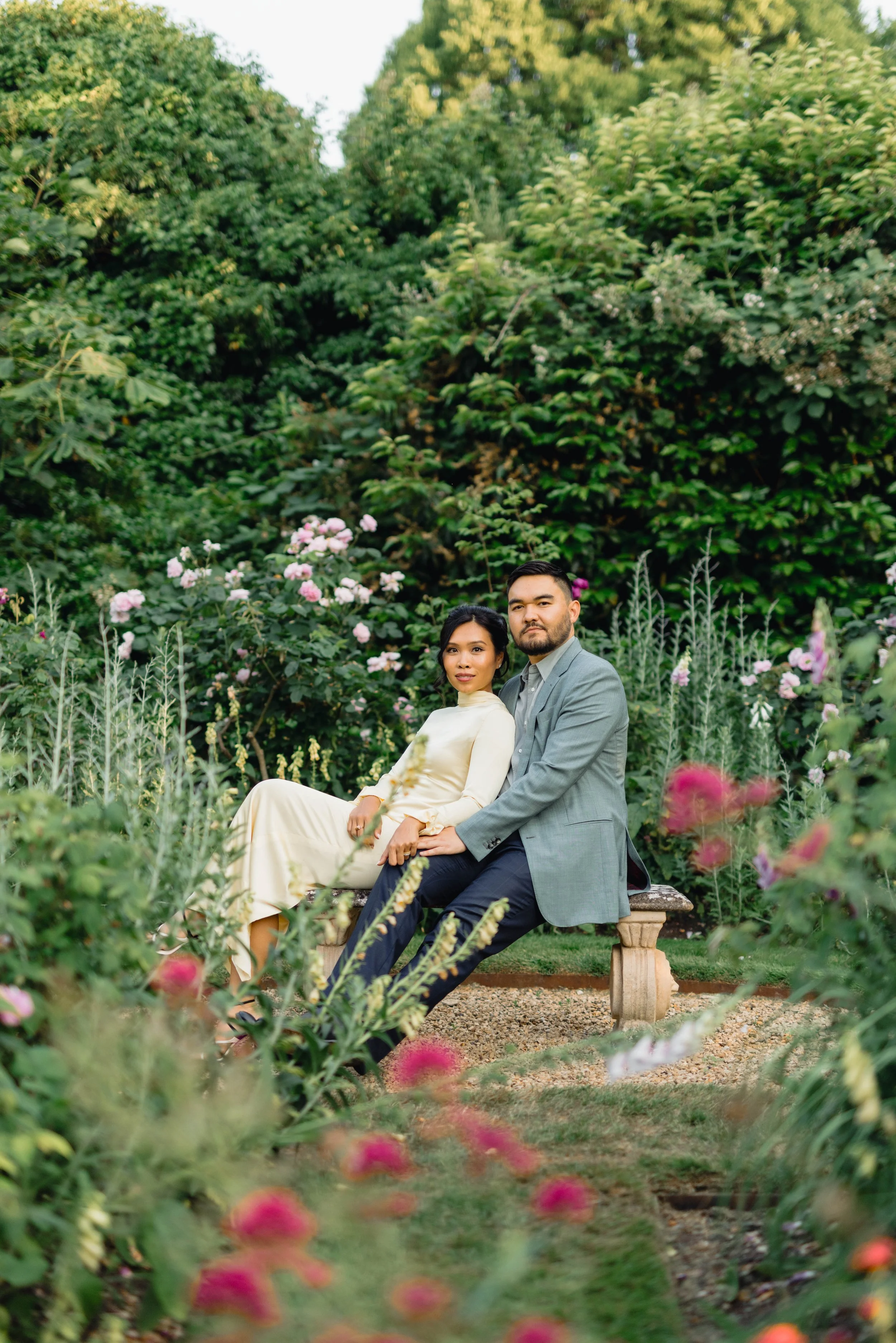A couple sitting on a stone bench in a lush garden with pink and white flowers and green foliage surrounding them.