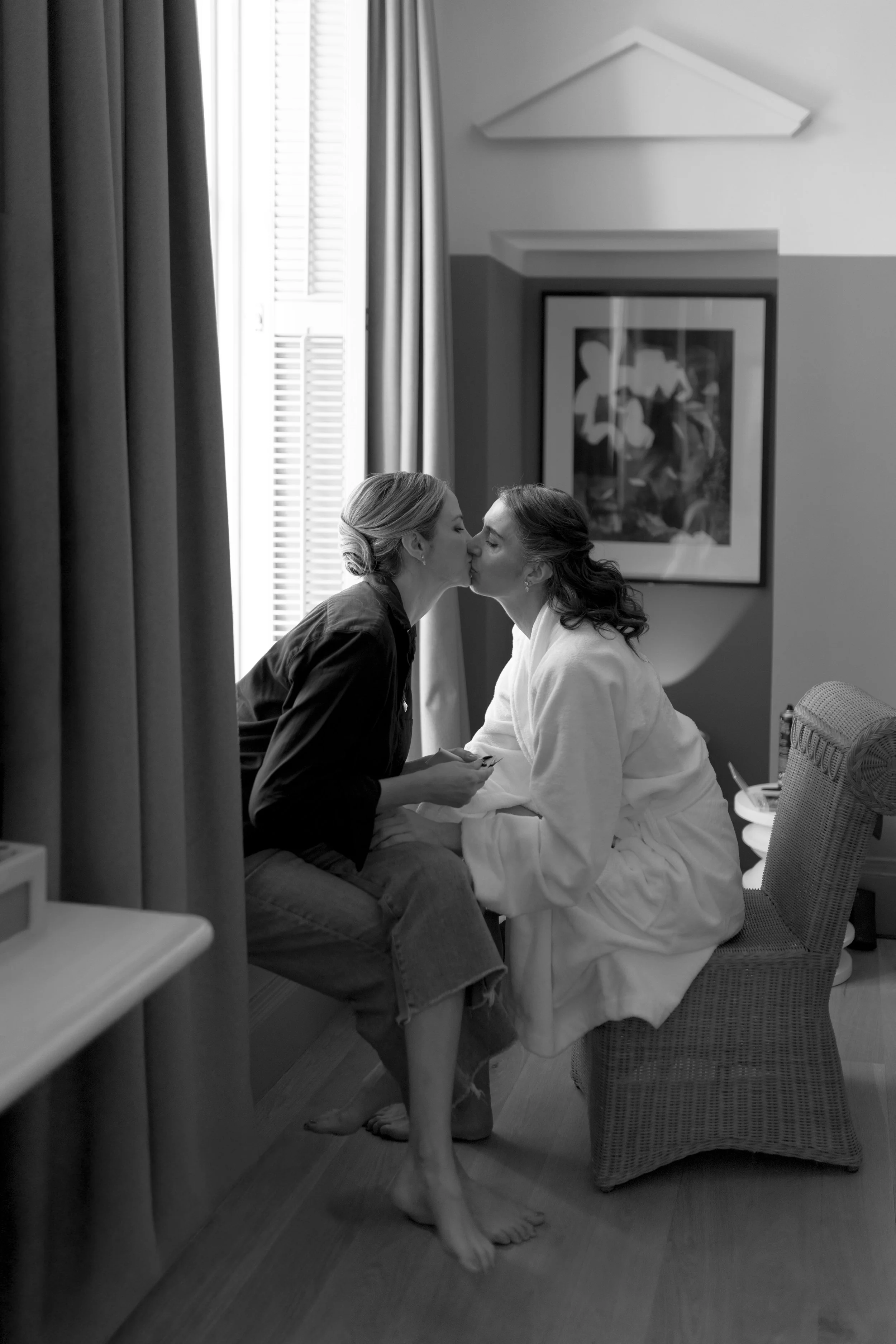 Two women kissing in a hotel room, one sitting on a bed and the other on a chair, near a window with closed blinds, in black and white.
