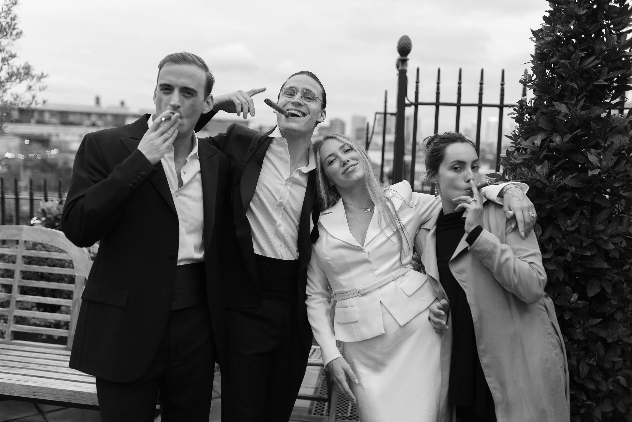 Four young adults in formal attire smoking cigars, standing on a rooftop with an urban cityscape in the background.