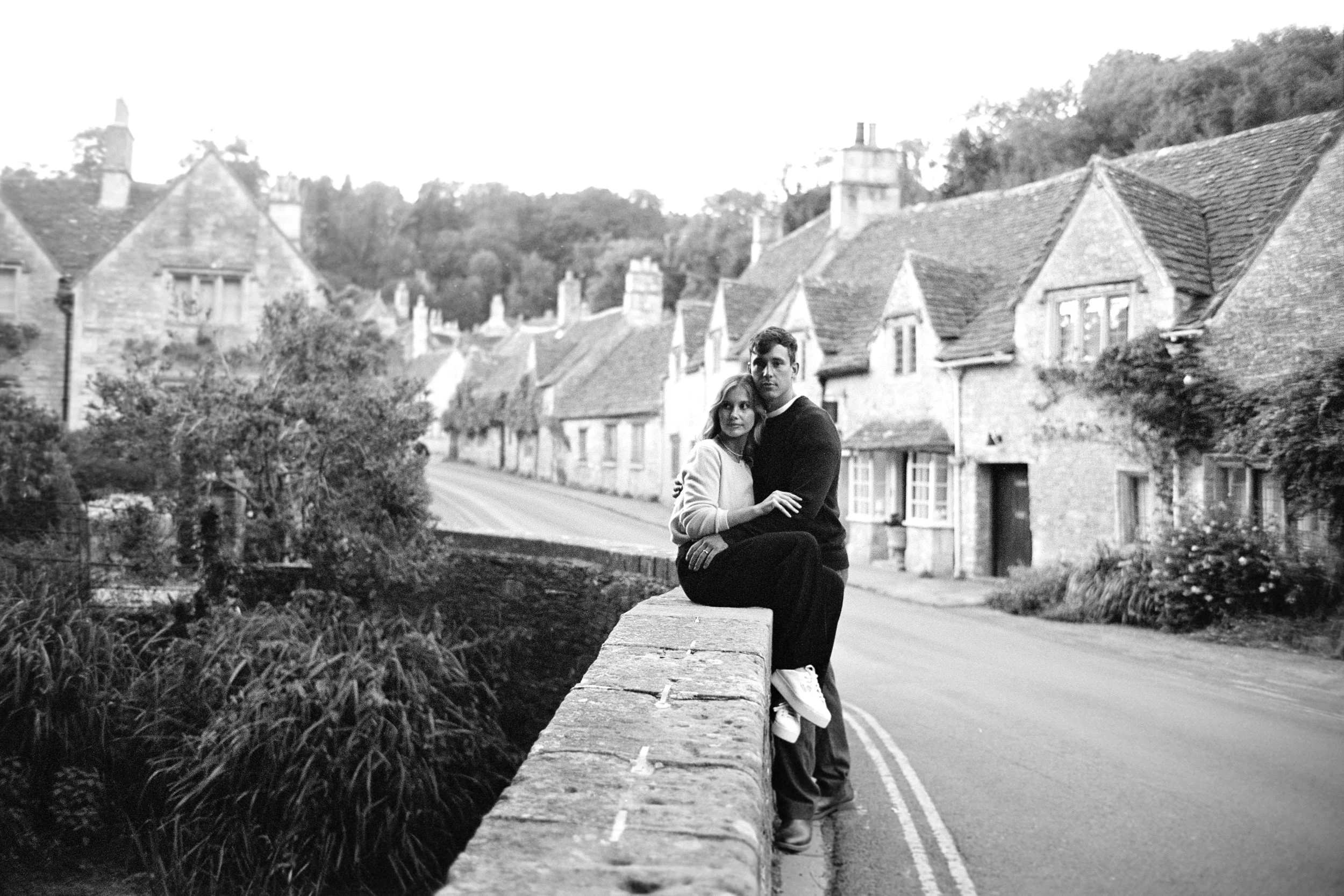 Black-and-white photo of a couple sitting on a low stone wall in a quaint village with stone cottages and lush trees in the background.