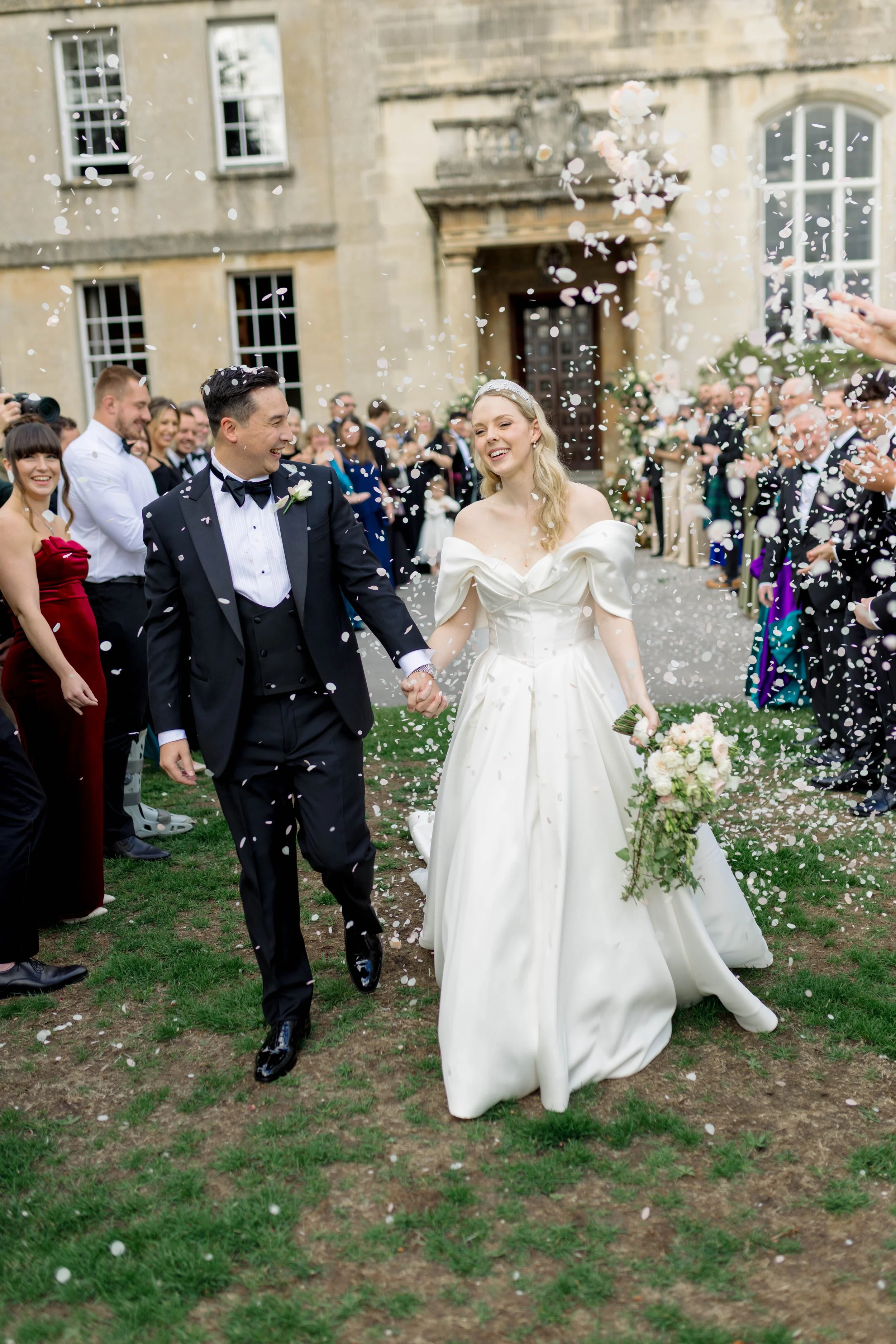 A bride and groom walking hand in hand, smiling, surrounded by guests throwing confetti, outside their Cotswold wedding venue, Elmore Court.