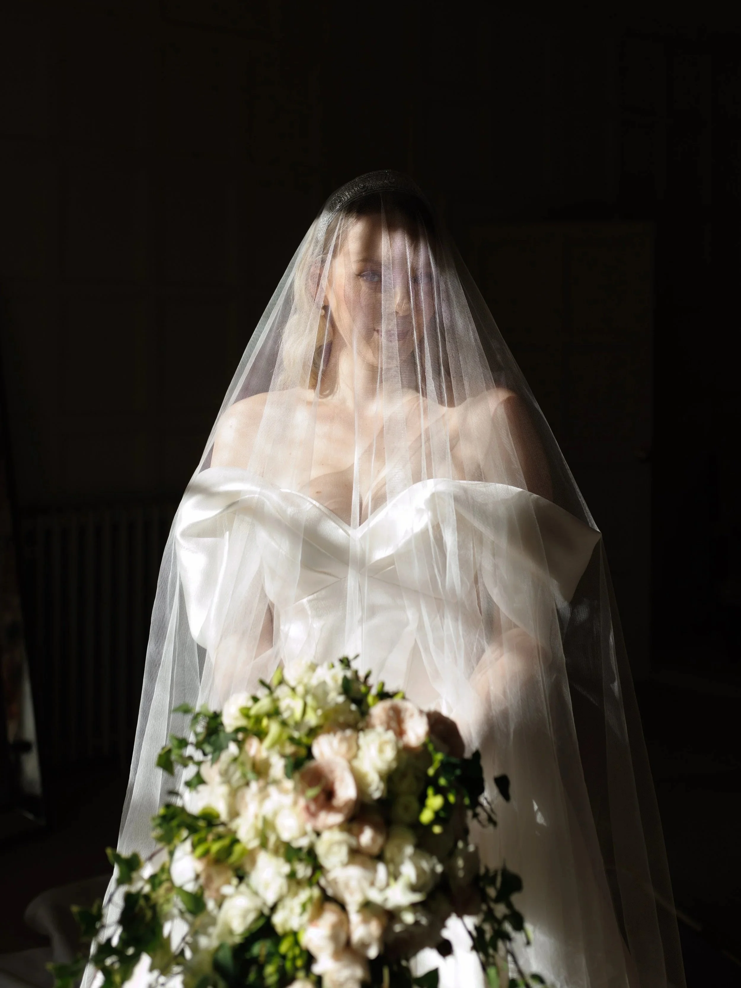 Bride in a wedding dress holding a bouquet, with a veil covering her face, standing in a dimly lit room.