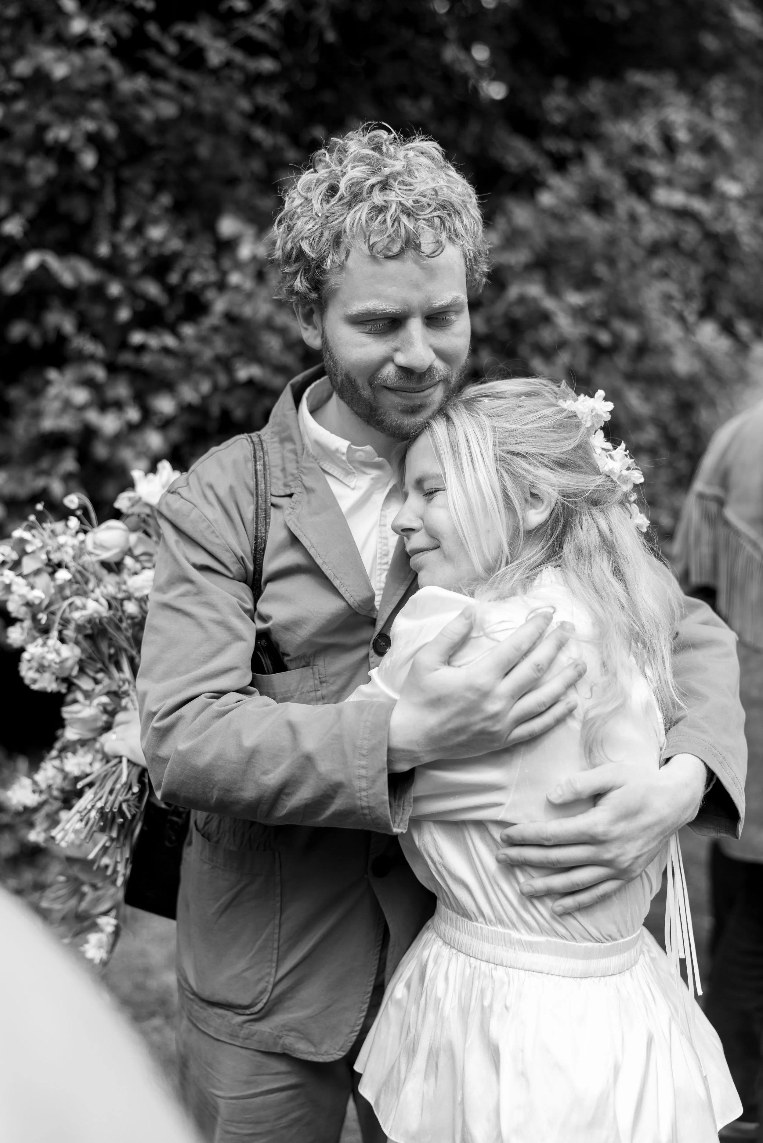 A man and woman embrace passionately outdoors, with the woman wearing a white dress and flowers in her hair, and the man holding her close against a background of trees.