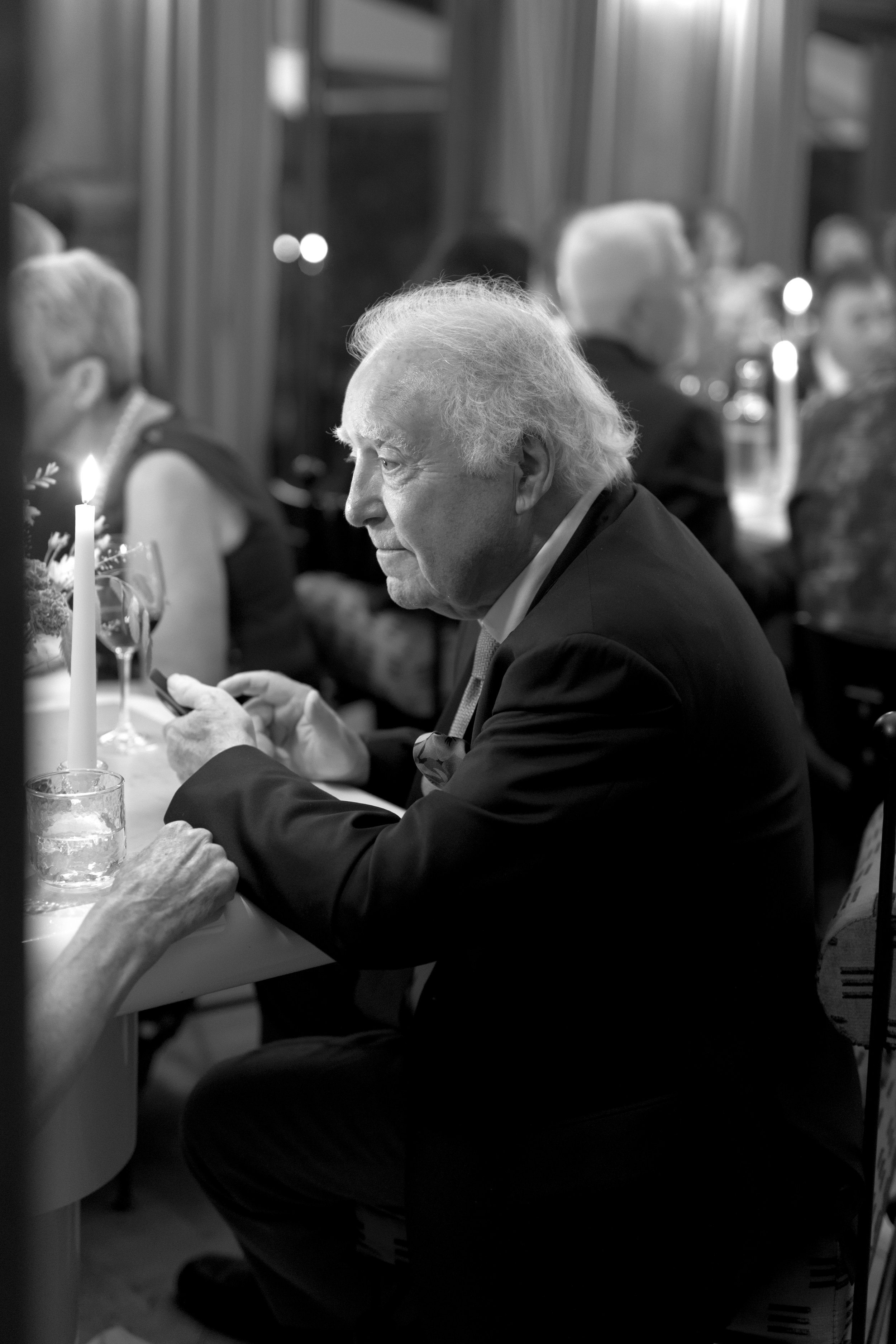 An elderly man with gray hair in a suit is sitting at a dinner table at night, looking at his phone, with a lit candle, wine glasses, and other diners in the background.