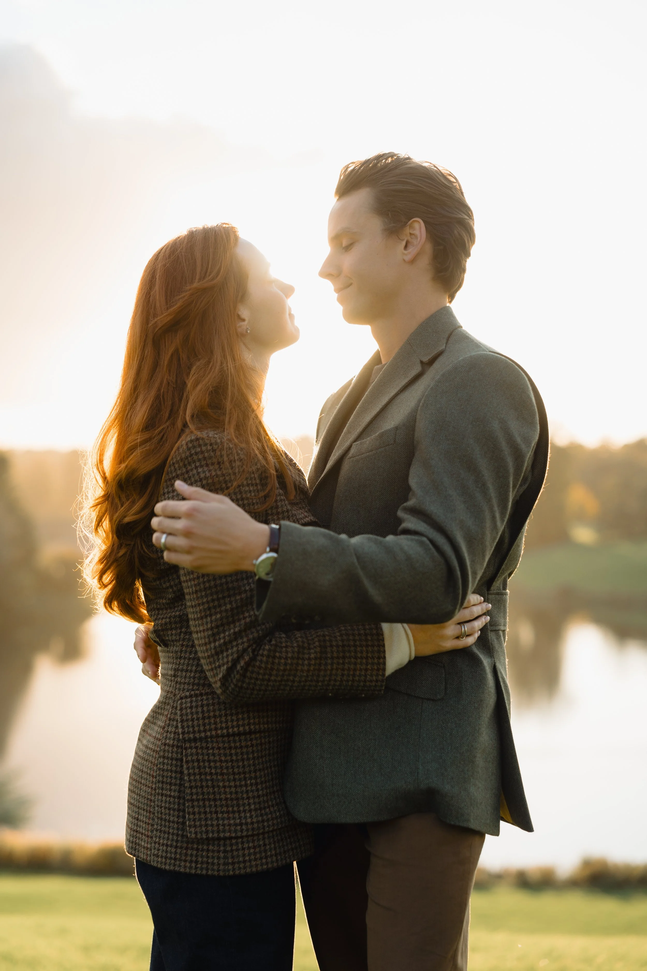 A man and woman stand close together outdoors, embracing each other as they look at one another with their eyes closed in a moment of intimacy during sunset.