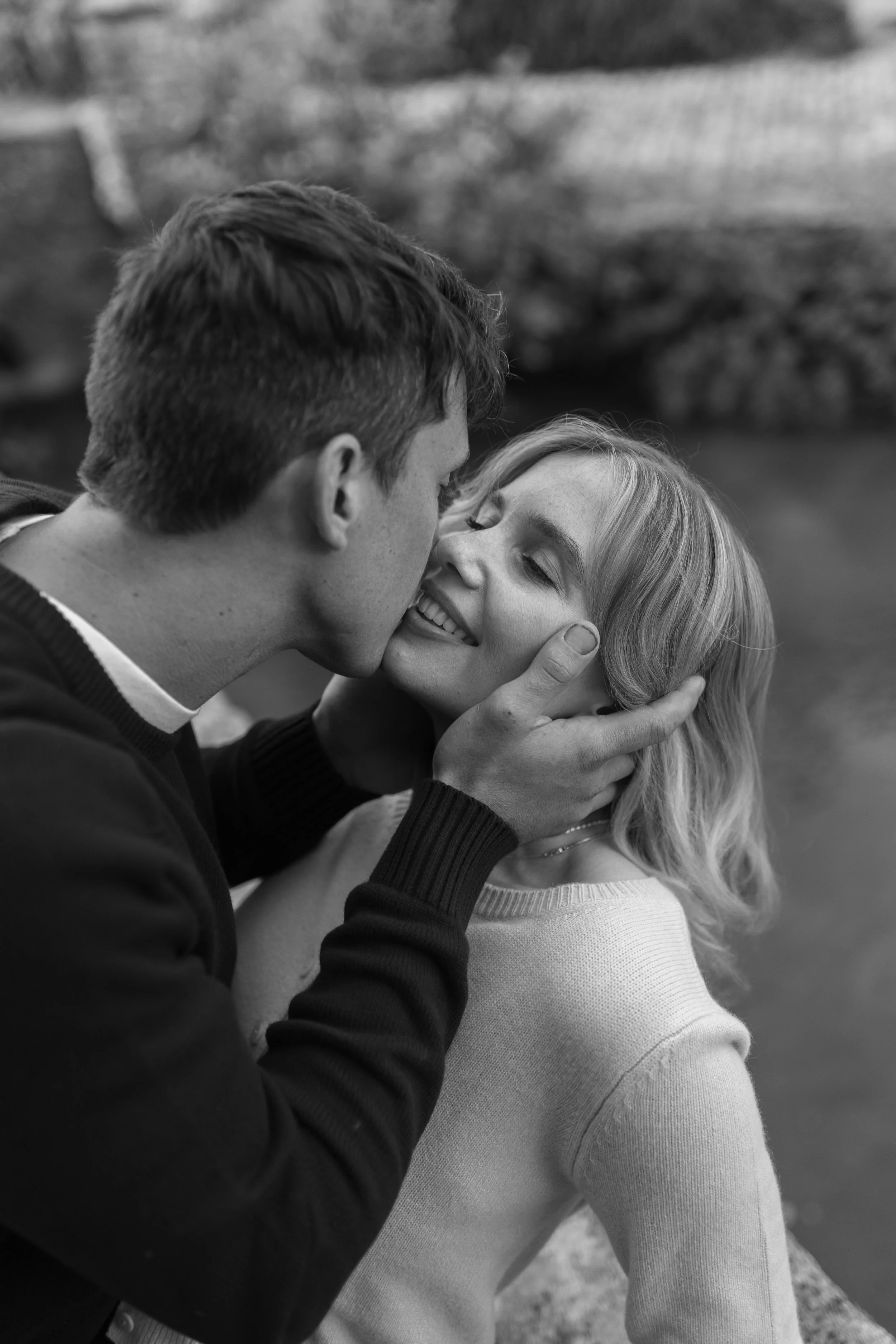 A black-and-white photo of a man and woman close together outdoors, with the man gently holding the woman's face as he kisses her on the lips. The woman is smiling and looking happy.