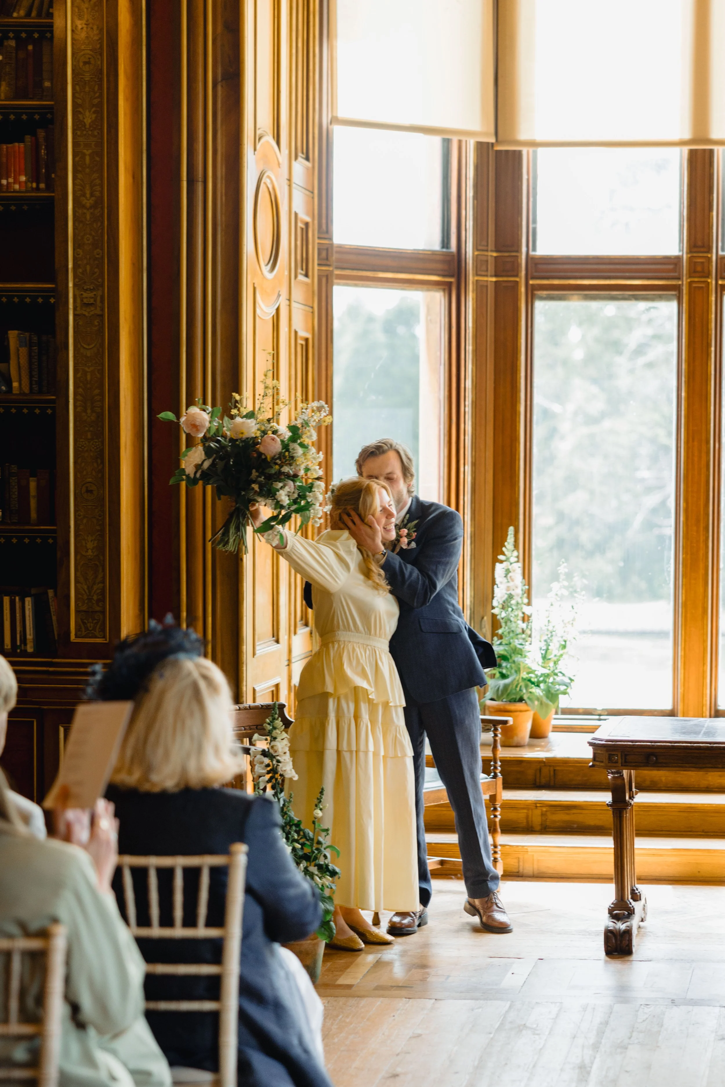 A couple is embracing during a wedding ceremony in a room with large wooden windows and bookshelves. The bride is wearing a cream-colored dress and holding a bouquet of flowers, while the groom is in a dark suit. Guests are seated and watching the mo