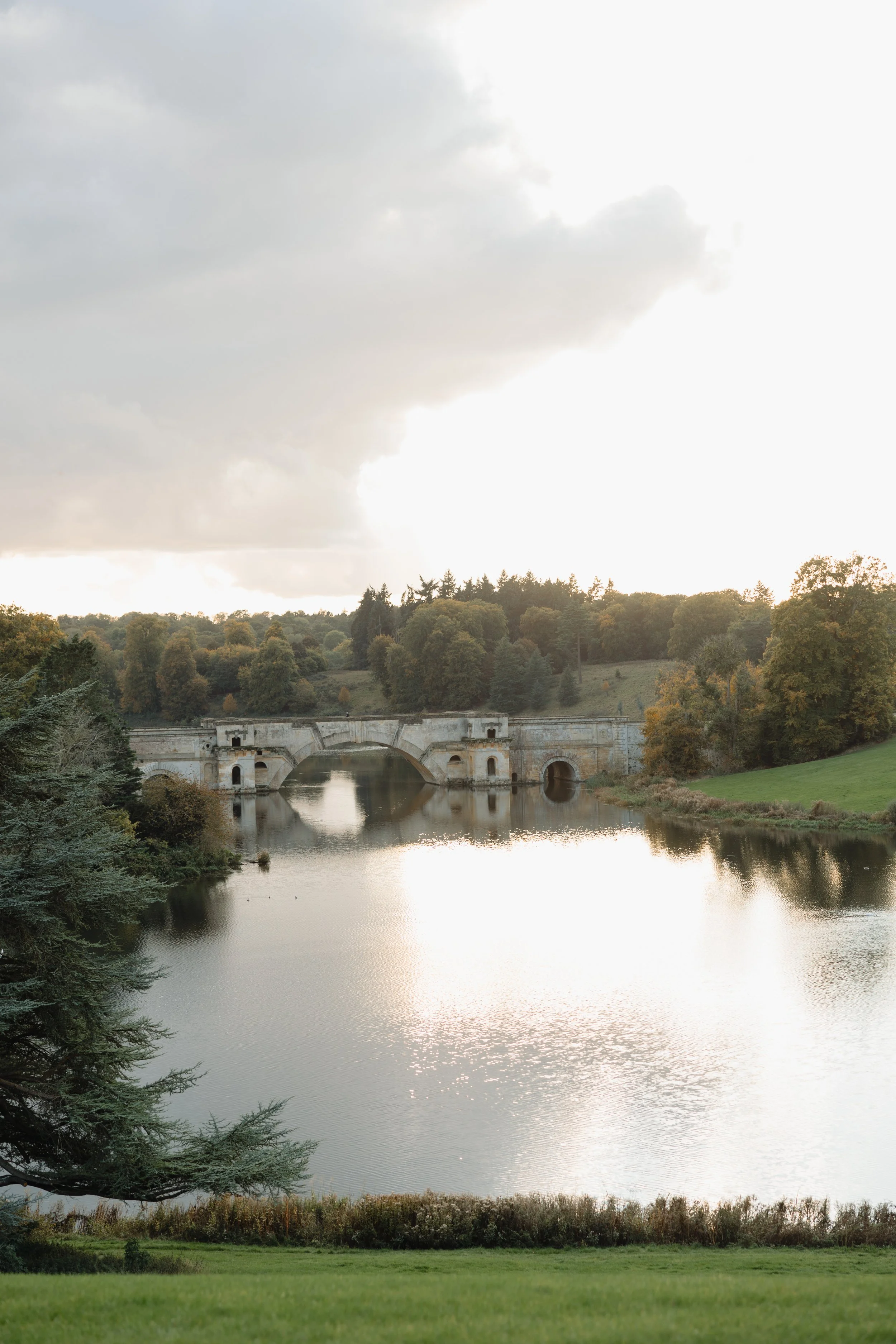 A calm river running through a green landscape with a stone bridge in the background, surrounded by trees and hills under a cloudy sky.