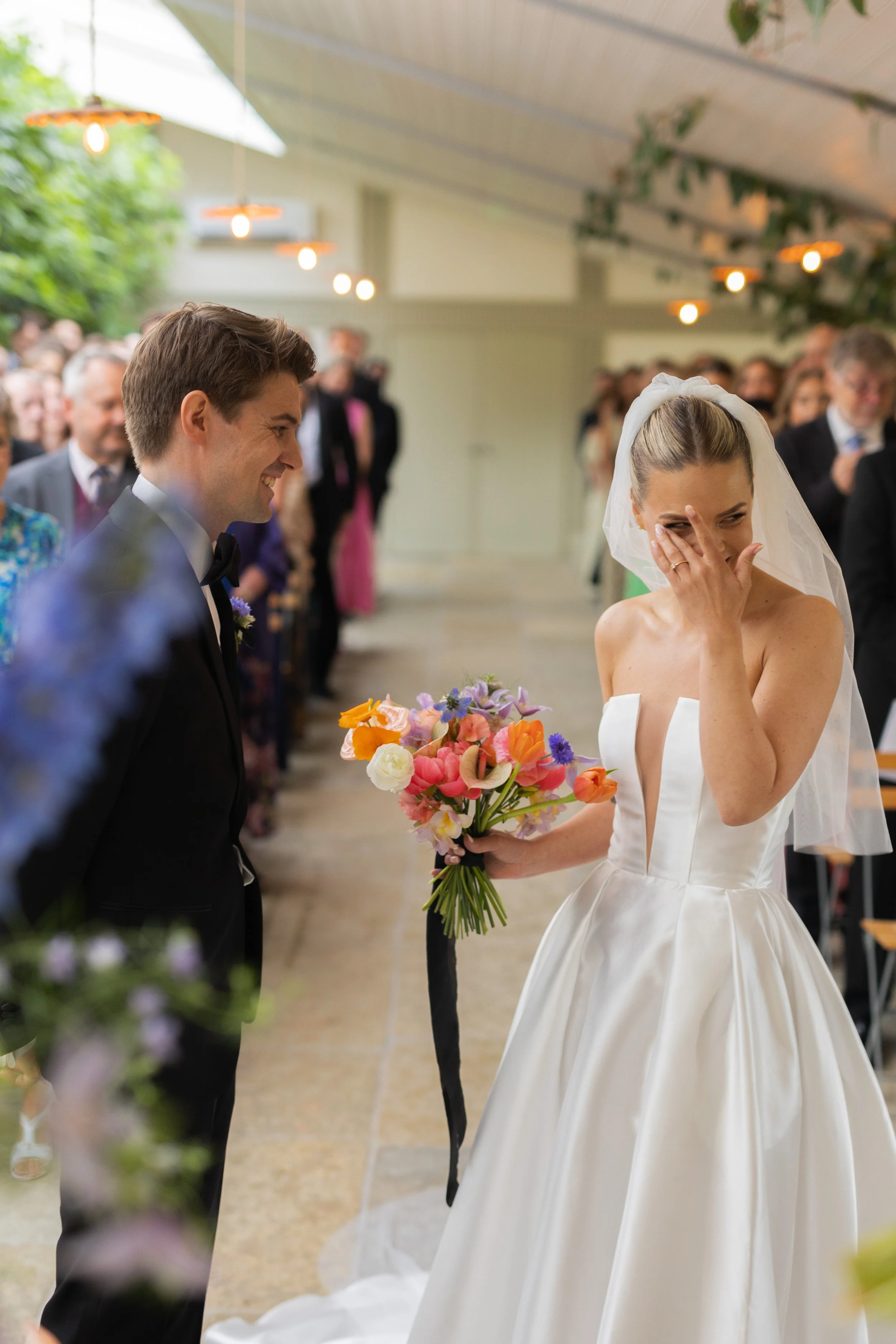 Bride and groom exchanging vows at wedding ceremony, bride covering her face and holding a colorful bouquet, groom smiling, guests seated and standing in the background.