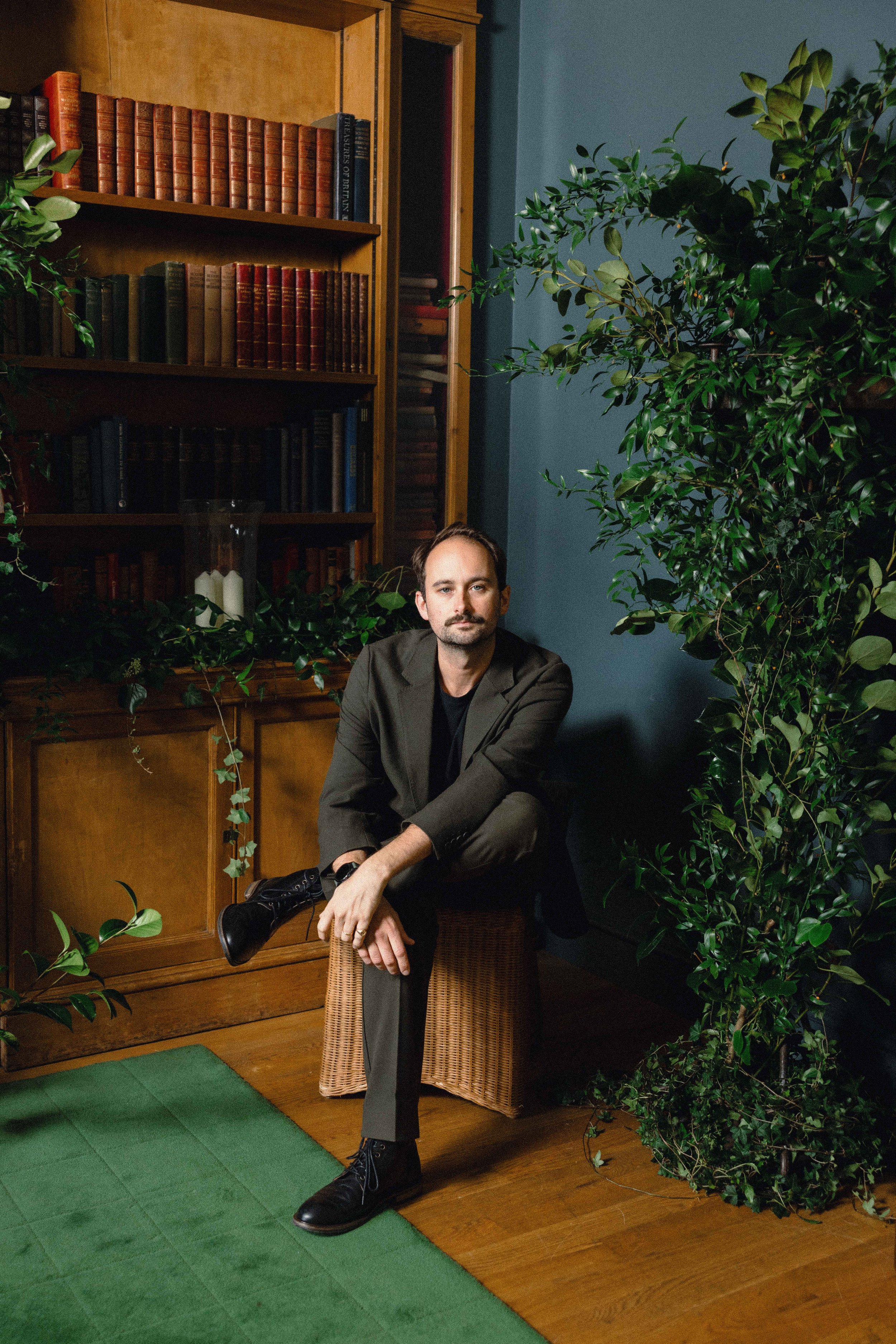 A man with a beard and mustache sitting on a wicker stool in a room with wooden flooring, surrounded by green plants and dark blue walls. There is a wooden bookshelf filled with books behind him.