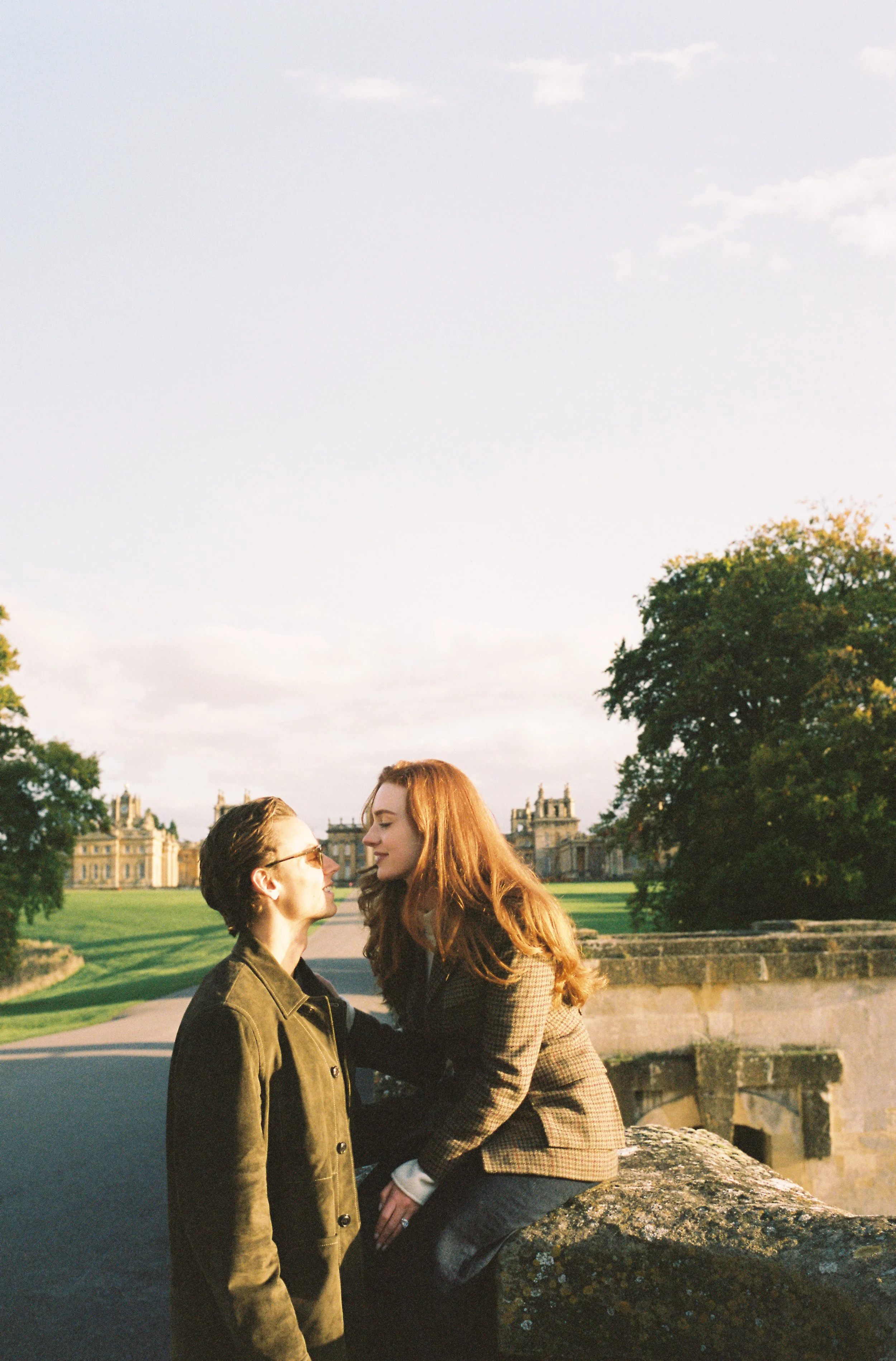 Two women looking at each other near a castle, with green lawns and trees in the background during sunset.