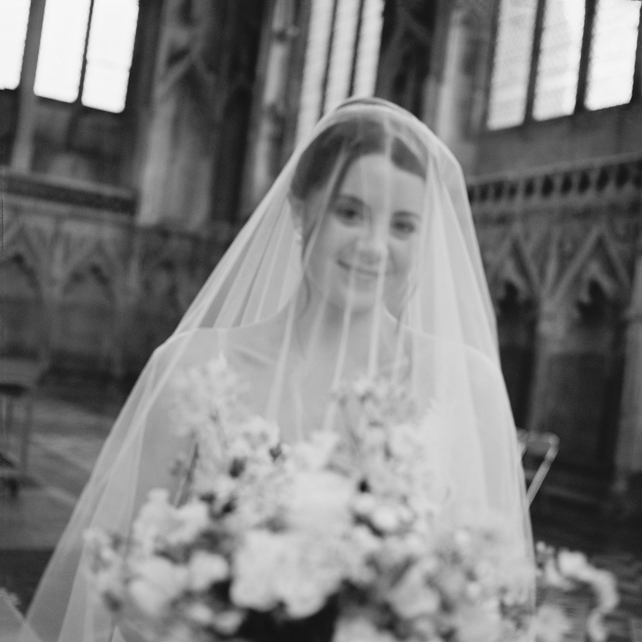 A bride smiling behind a sheer veil, holding a floral bouquet, inside Ely Cathedral with high stained glass windows.