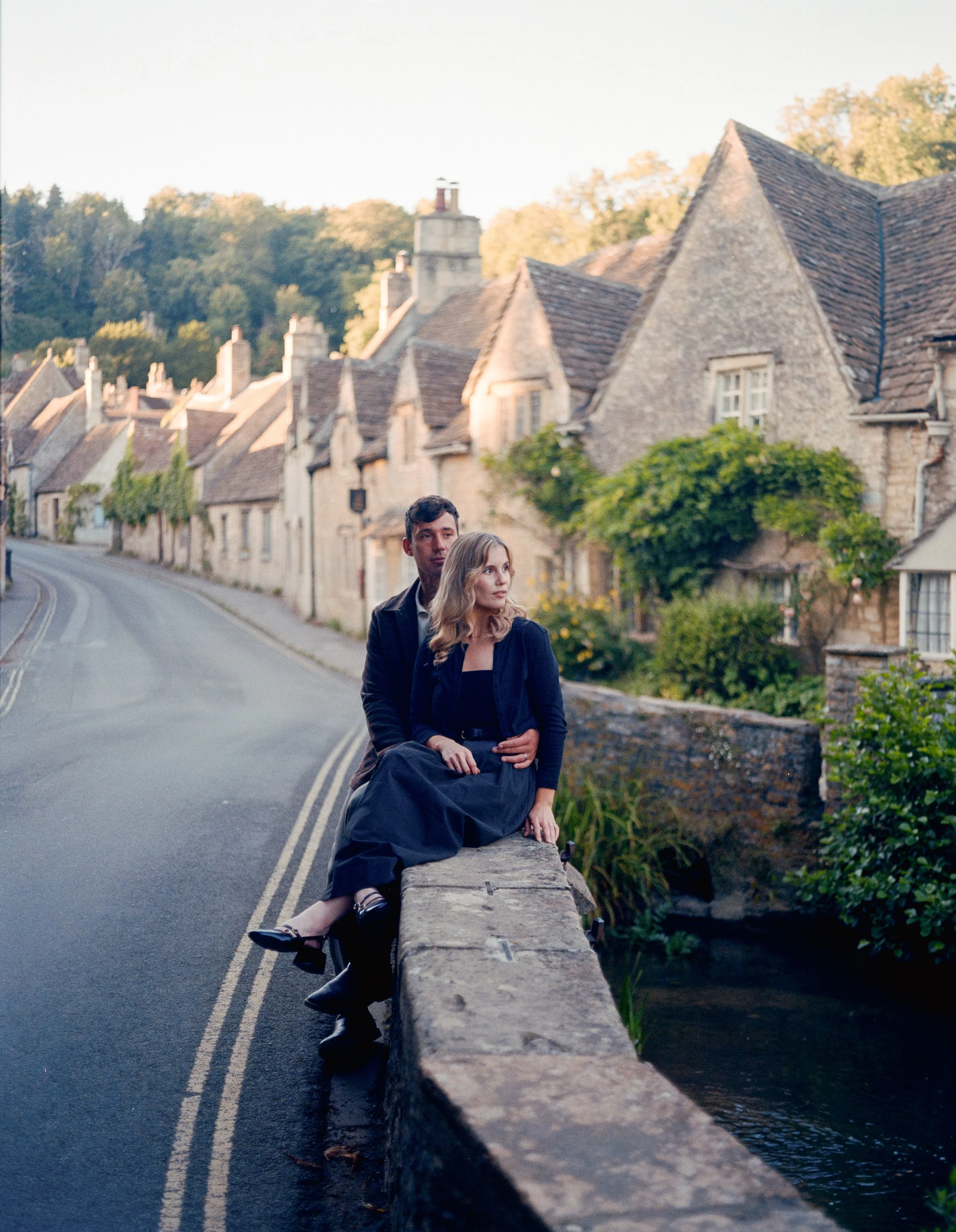A man and woman sitting on a stone wall beside a small stream in a village with historic stone cottages, during the daytime.
