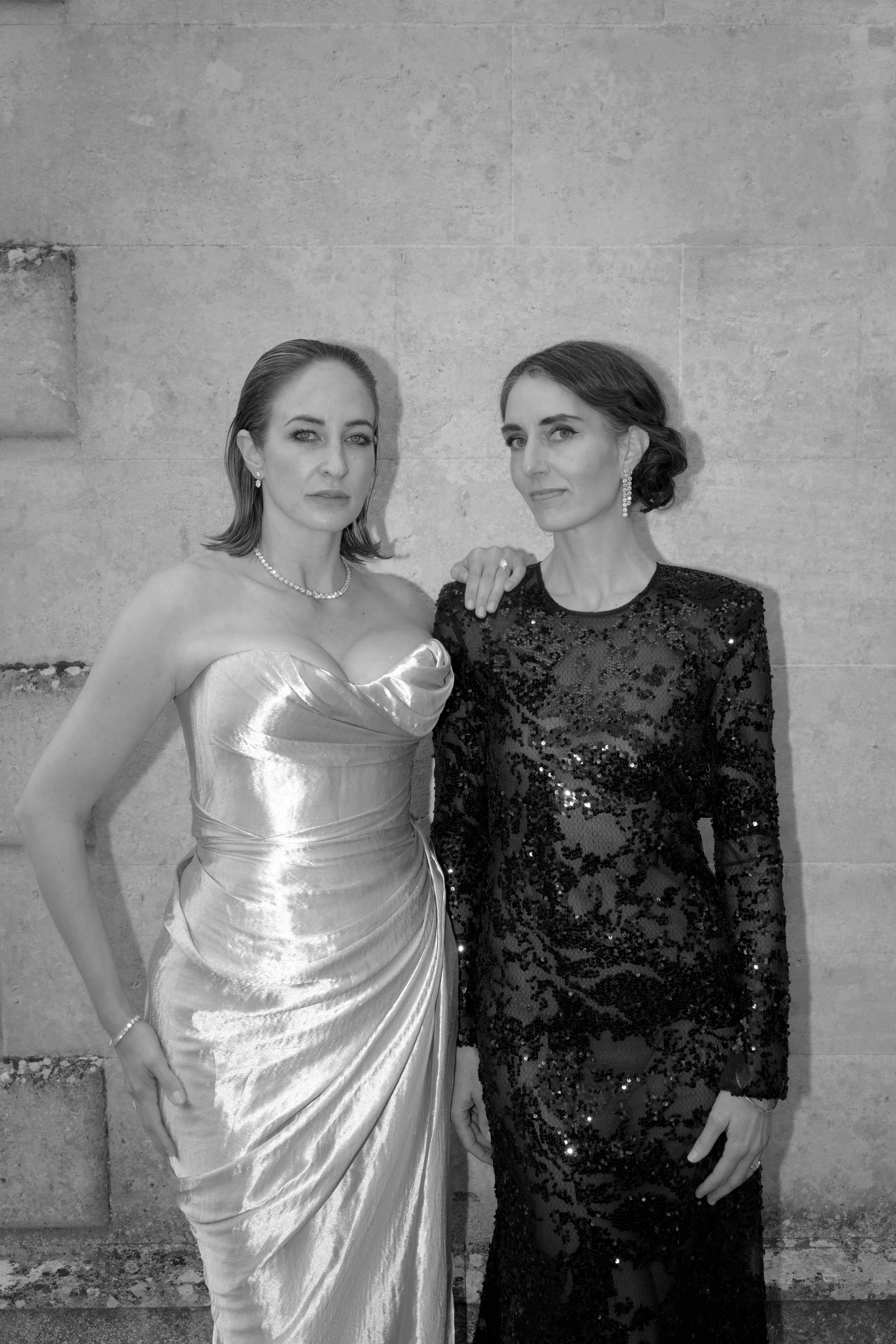 Two women dressed in elegant evening gowns pose together against a stone wall. One wears a shiny, strapless gown, while the other wears a black sequined dress. They both wear jewelry and have their hair styled formally.