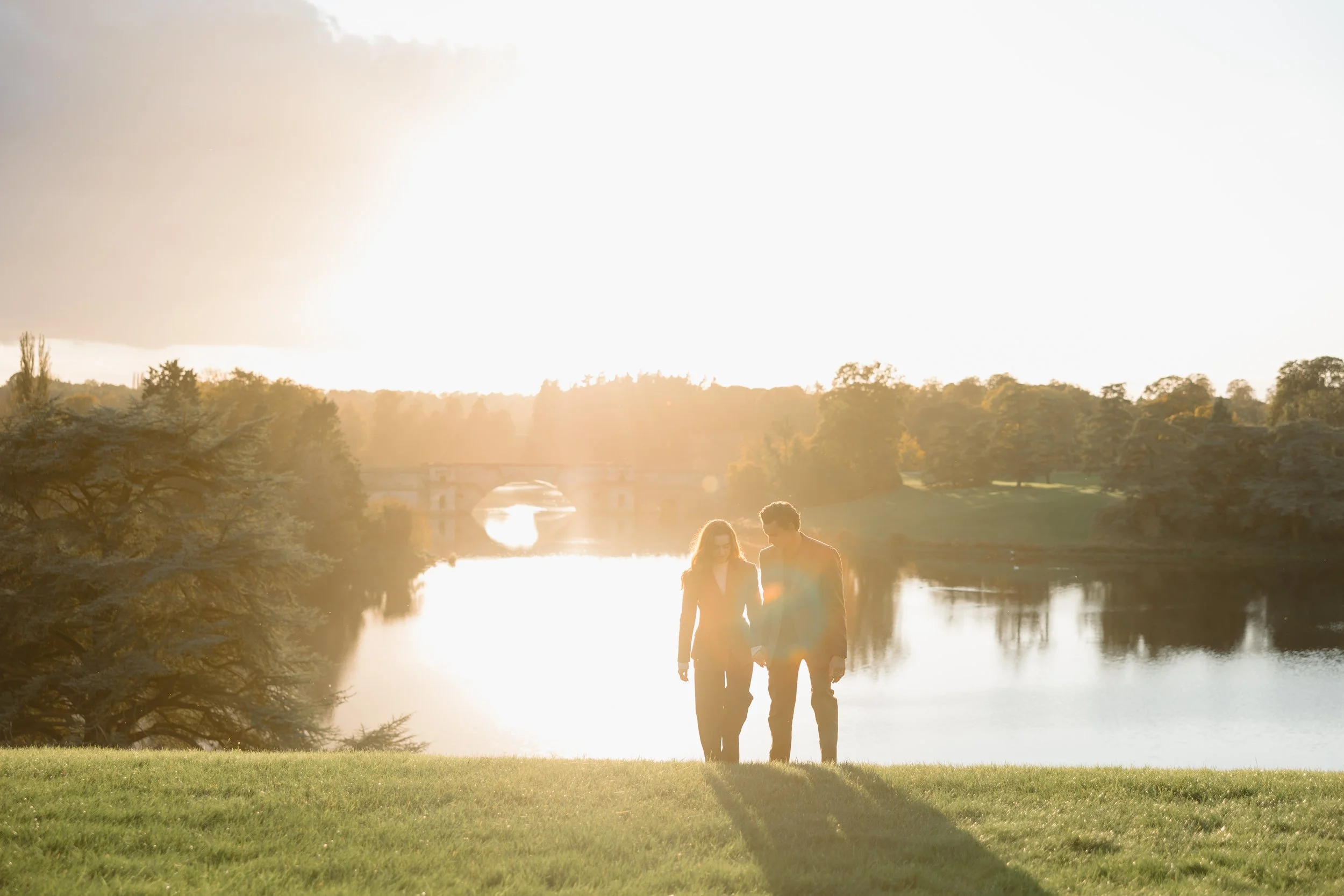 A couple walking hand in hand by a lake during sunset with trees and a bridge in the background.