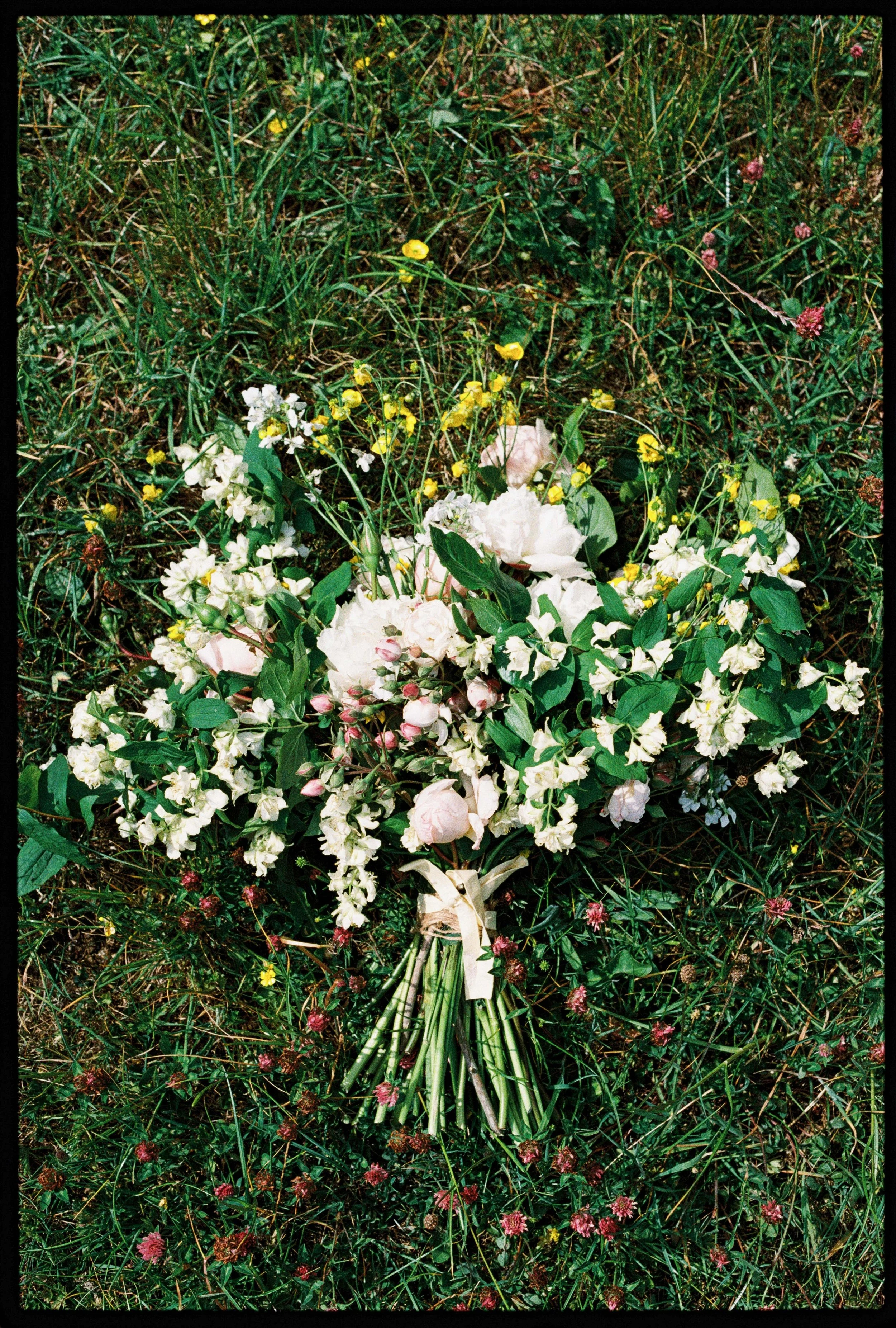 Bouquet of white and pink flowers lying on green grass with small pink and yellow flowers surrounding it