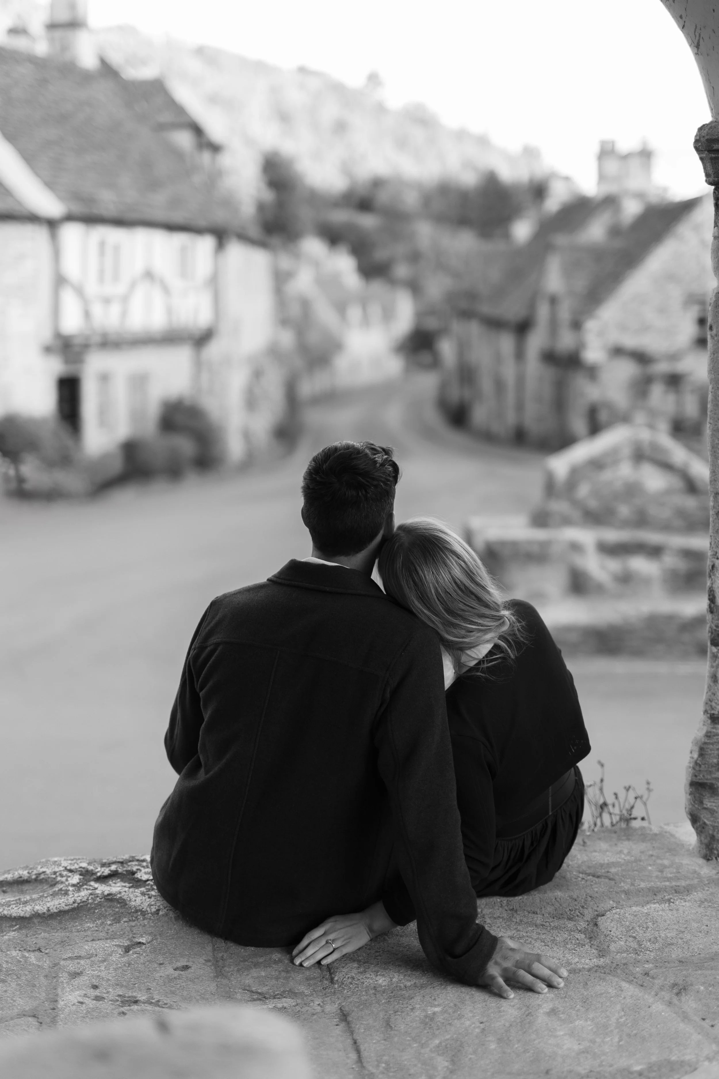 A couple sitting on a ledge looking out over a blurred village scene in black and white.