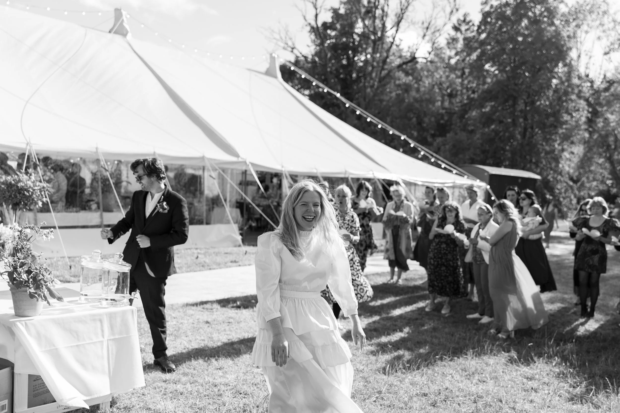 Black and white photo of a group of people at an outdoor gathering or party. A woman in a white dress is smiling and walking towards the camera, with a large tent and trees in the background.