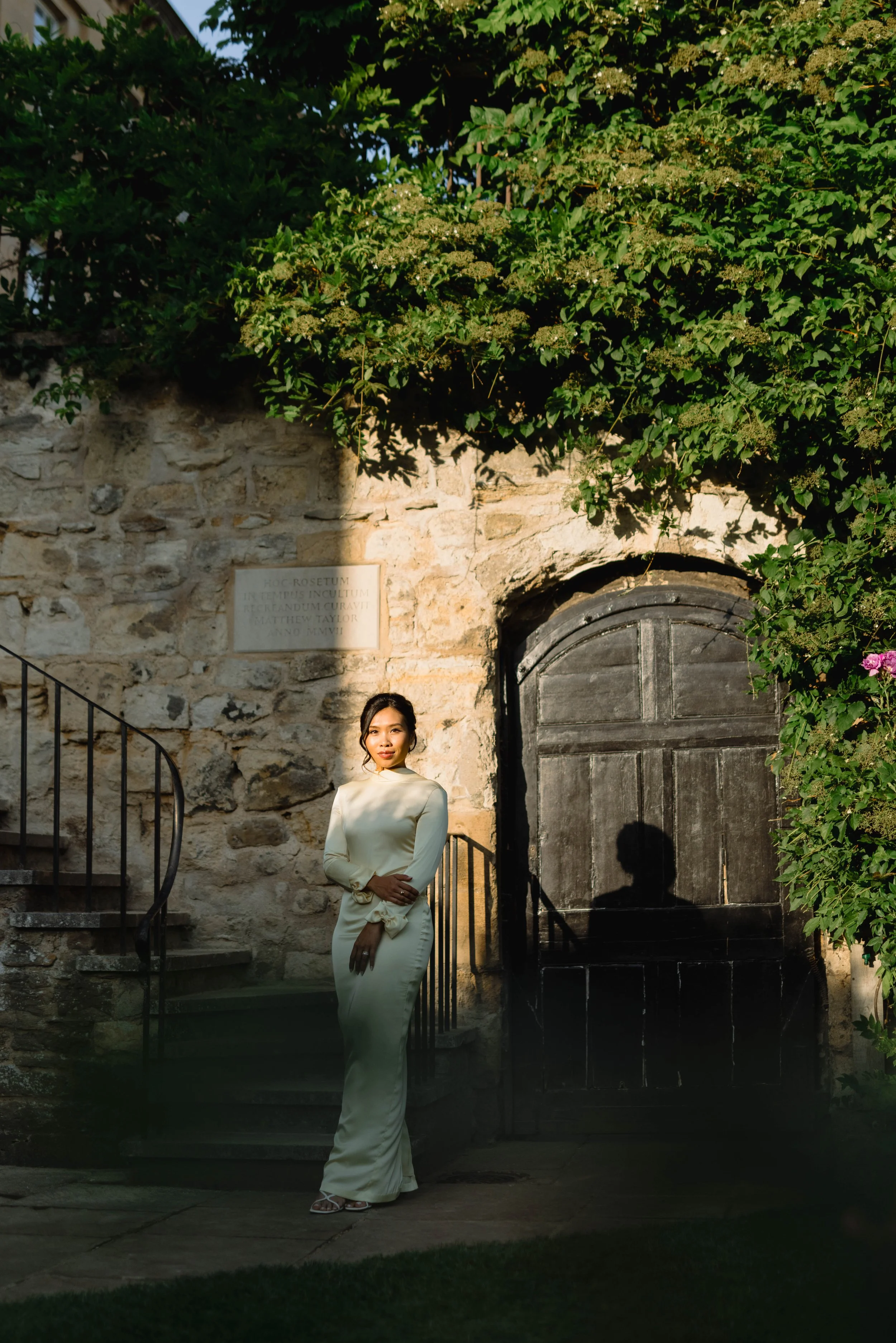 A woman in a long, light-colored dress standing outdoors near an old stone building with a dark wooden door, surrounded by lush green foliage. Her shadow is cast on the door, and the scene is bathed in warm sunlight.