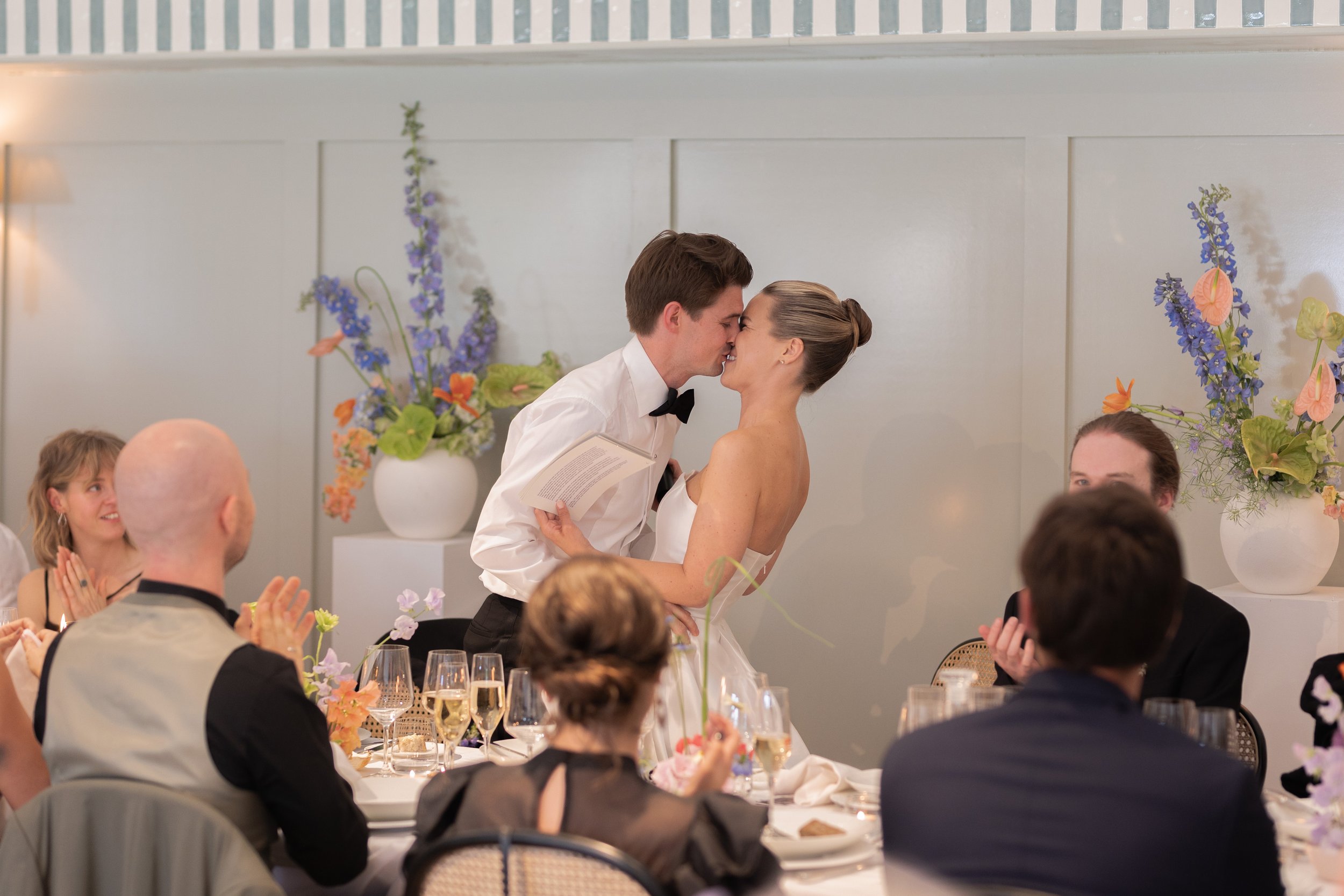 A wedding reception scene where a bride and groom are kissing at their wedding ceremony surrounded by friends and family at a decorated table with floral arrangements and champagne glasses.