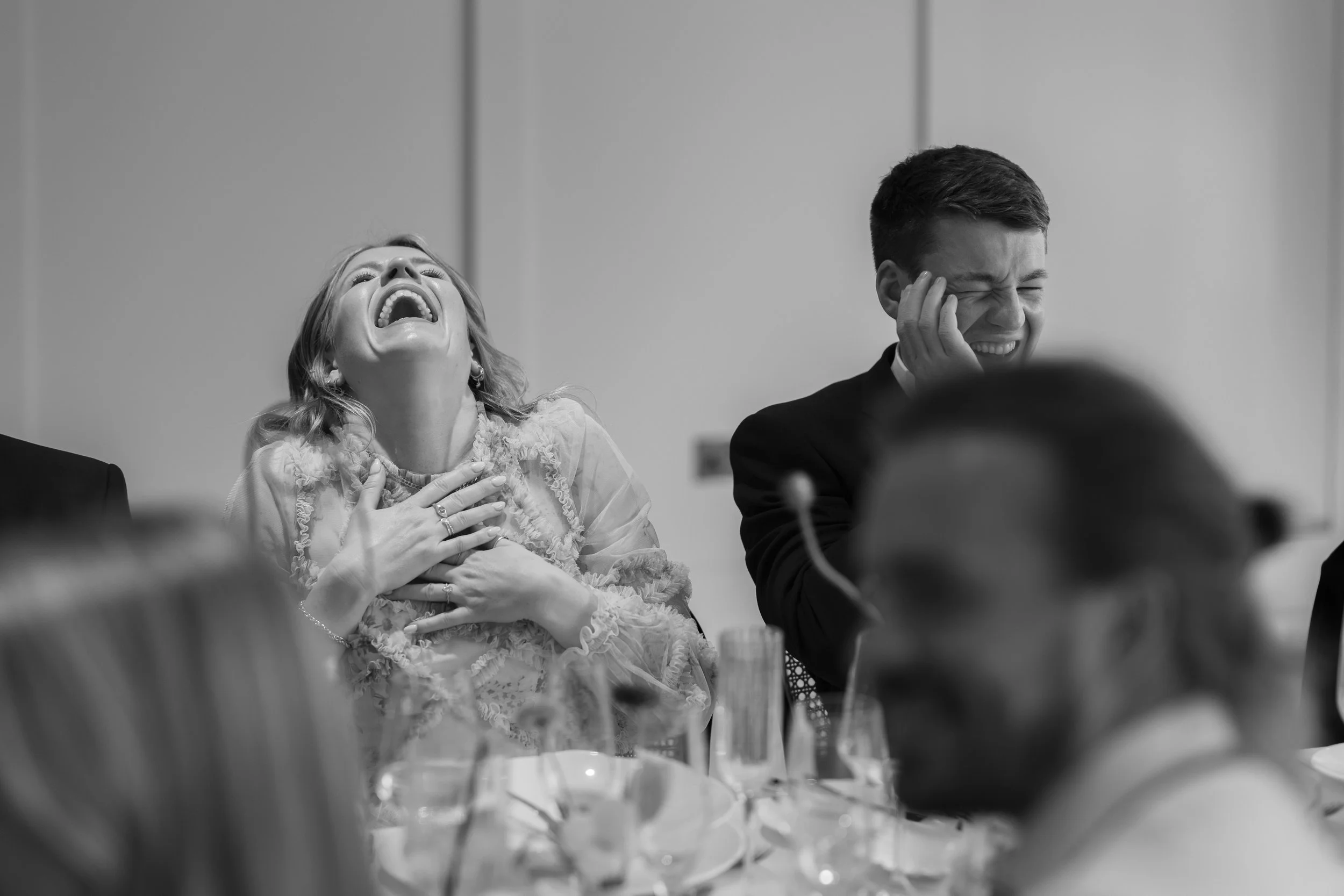 Black and white photo of two people, a woman and a man, laughing and crying at a formal event or celebration, sitting at a table with wine glasses and tableware.