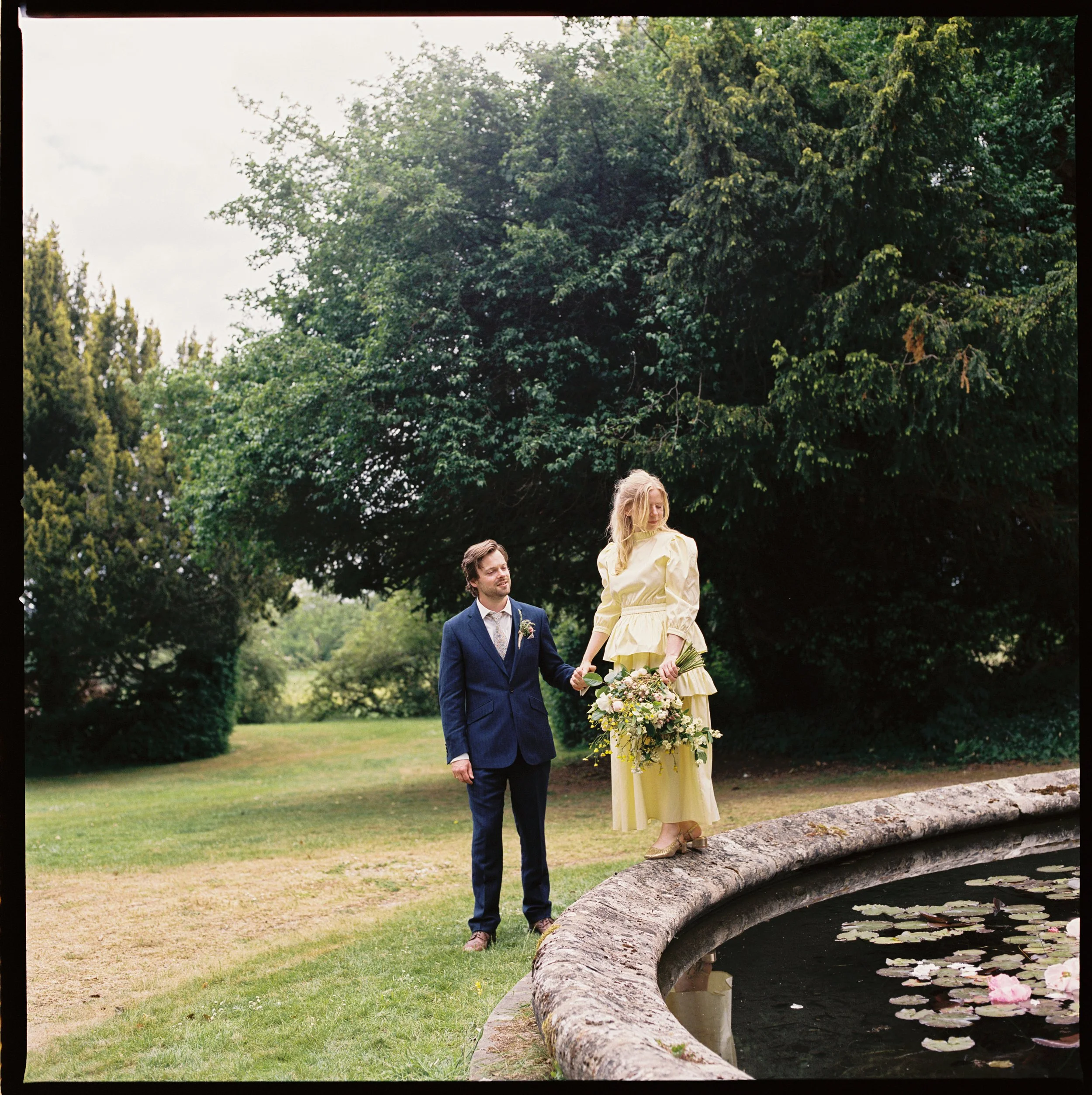 A bride standing on a stone edge next to a pond with water lilies, holding a bouquet, and a groom in a blue suit holding her hand, outdoors in a lush green park.