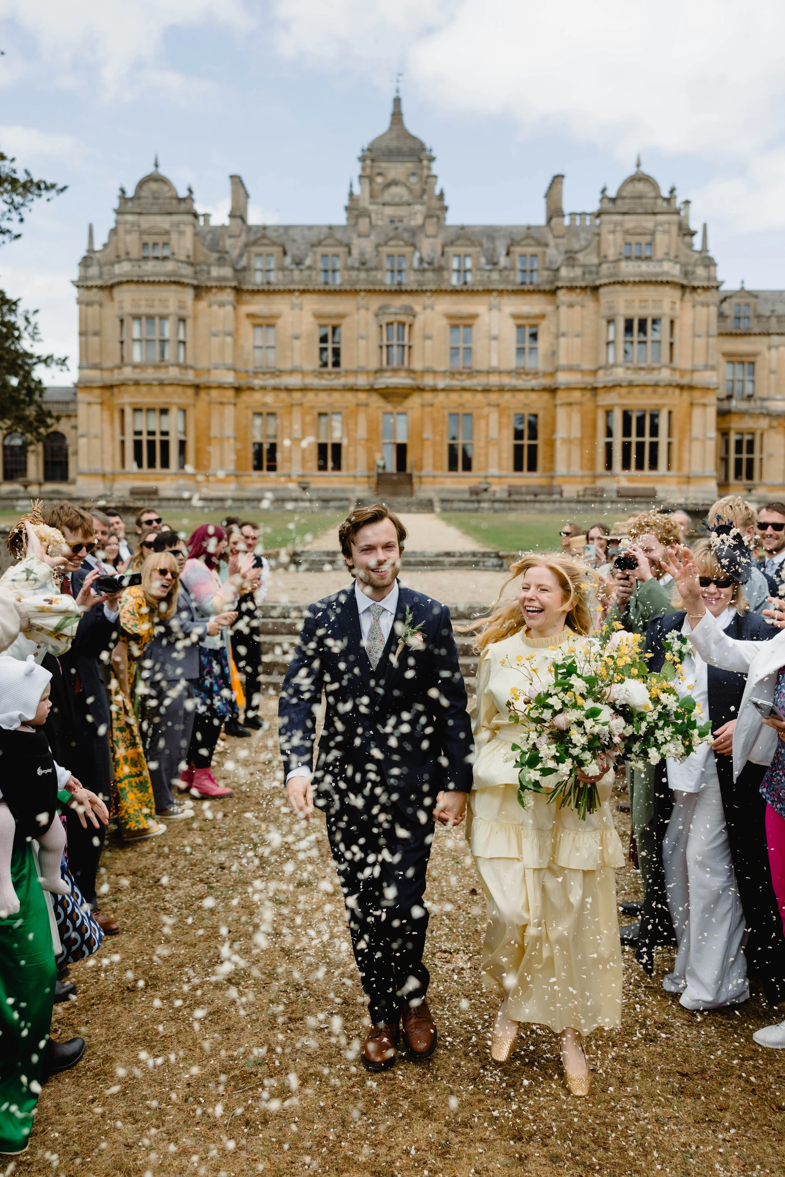 A bride and groom walking hand in hand outside a historic building, surrounded by congratulating friends and family, with confetti flying in the air during a wedding celebration.