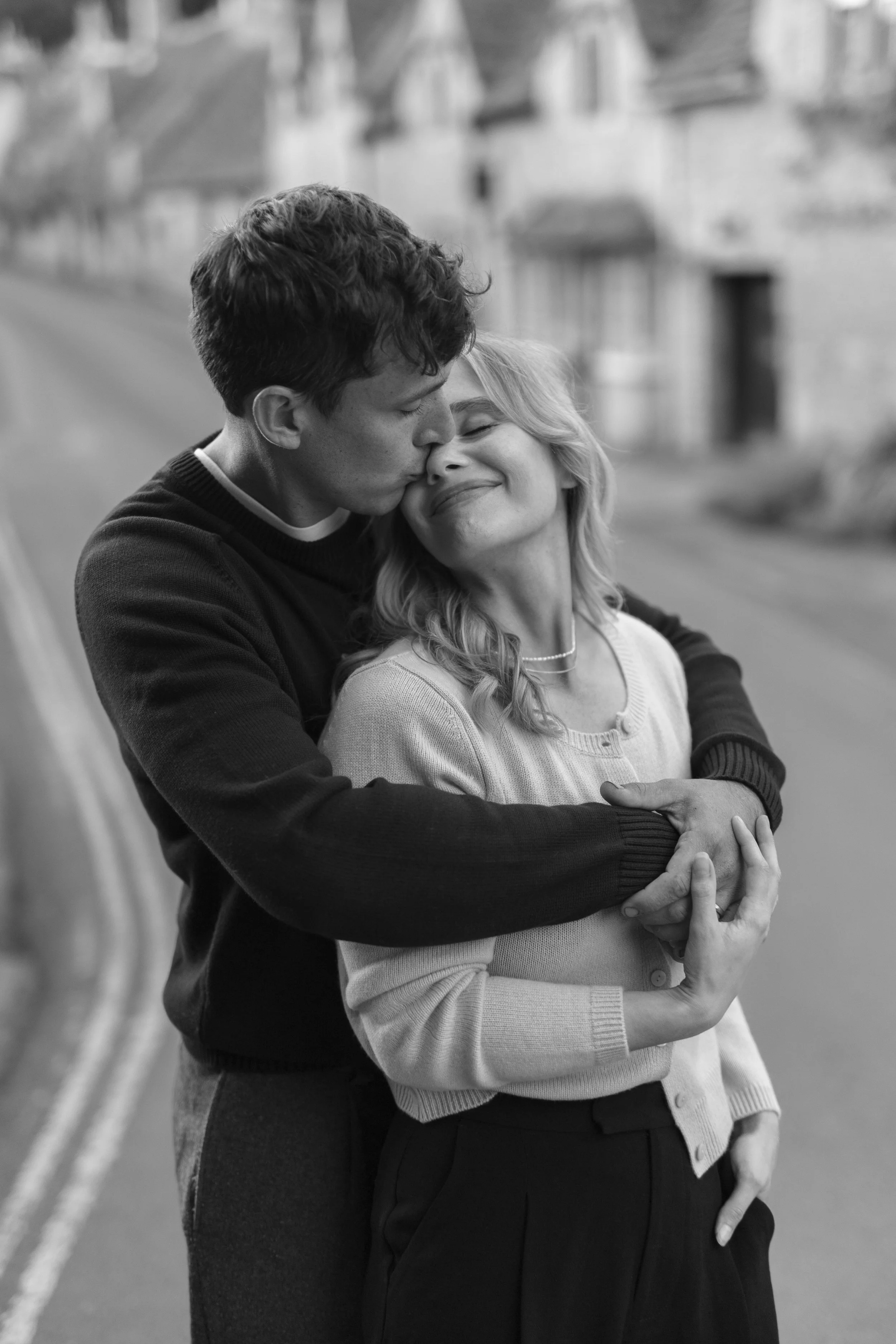 A black-and-white photo of a young man kissing an older woman on the forehead as they embrace on a street.
