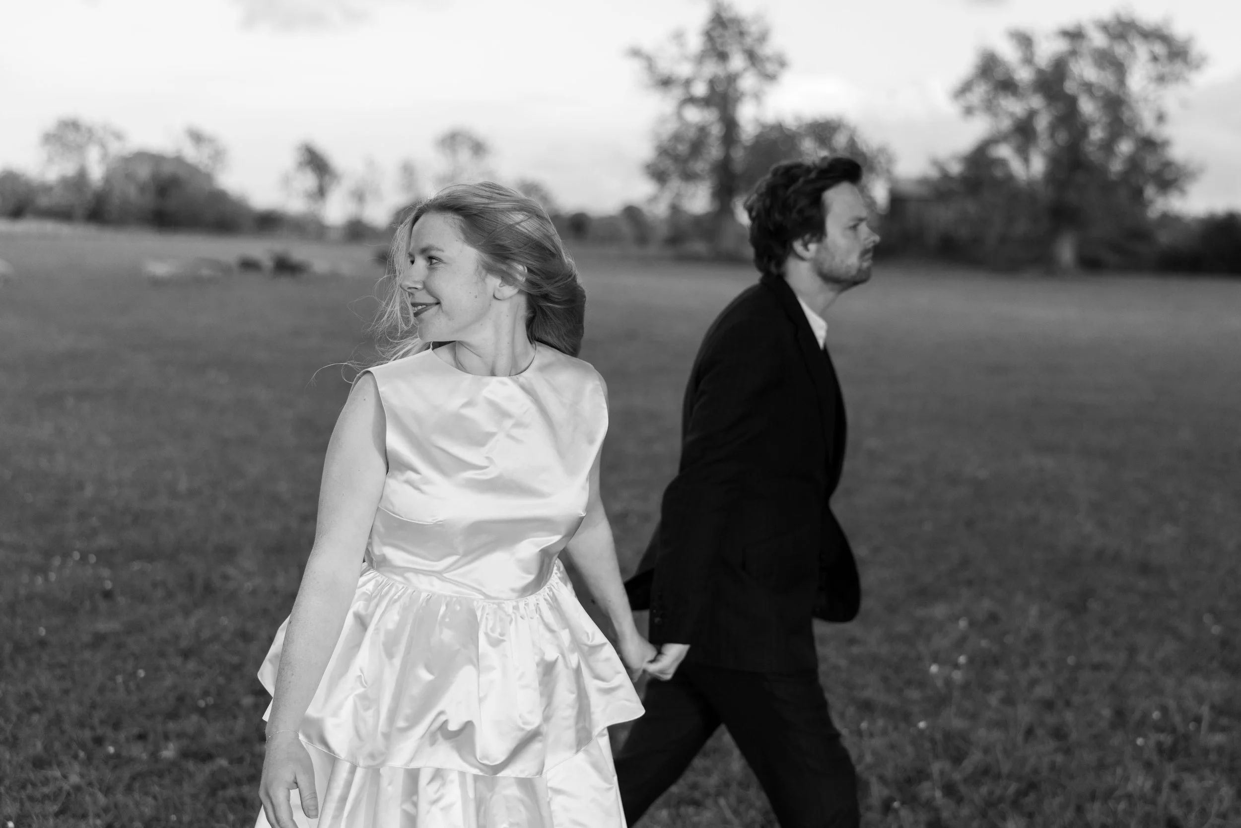 A black-and-white photo of a man and a woman holding hands and walking outdoors in a grassy field with trees in the background. The woman is smiling and looking to the left, wearing a satin dress, while the man looks to the right, wearing a dark suit