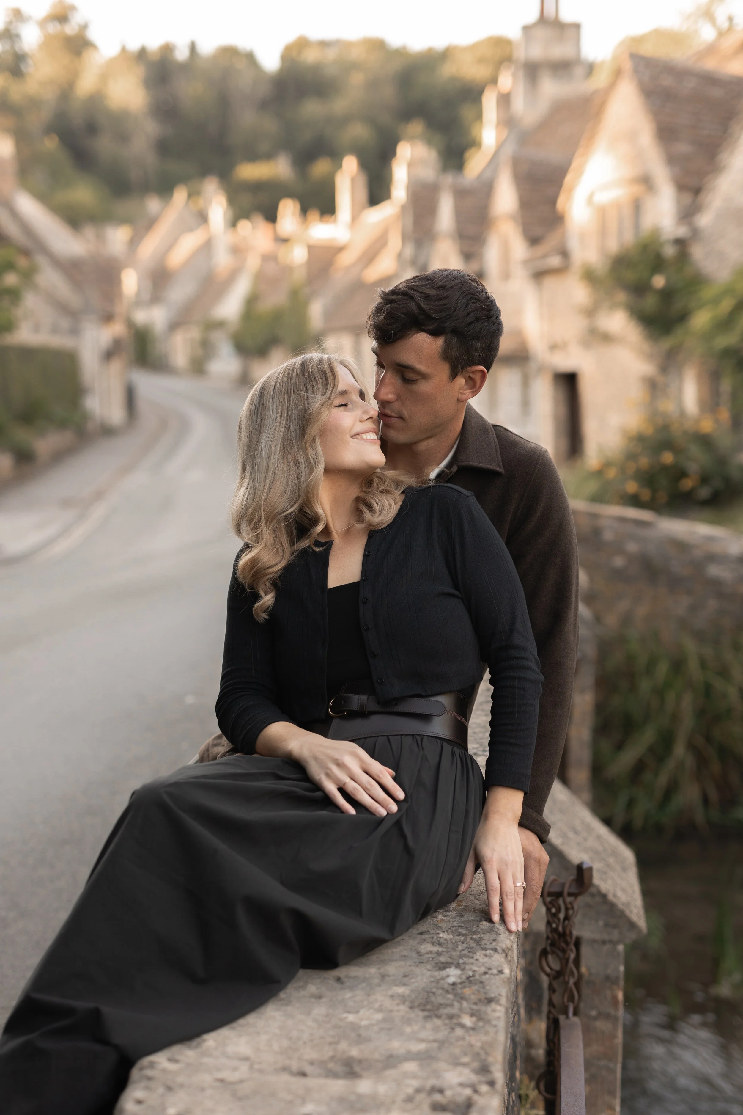 A couple embracing by a stone bridge on a quiet street with European-style houses in the background during sunset.