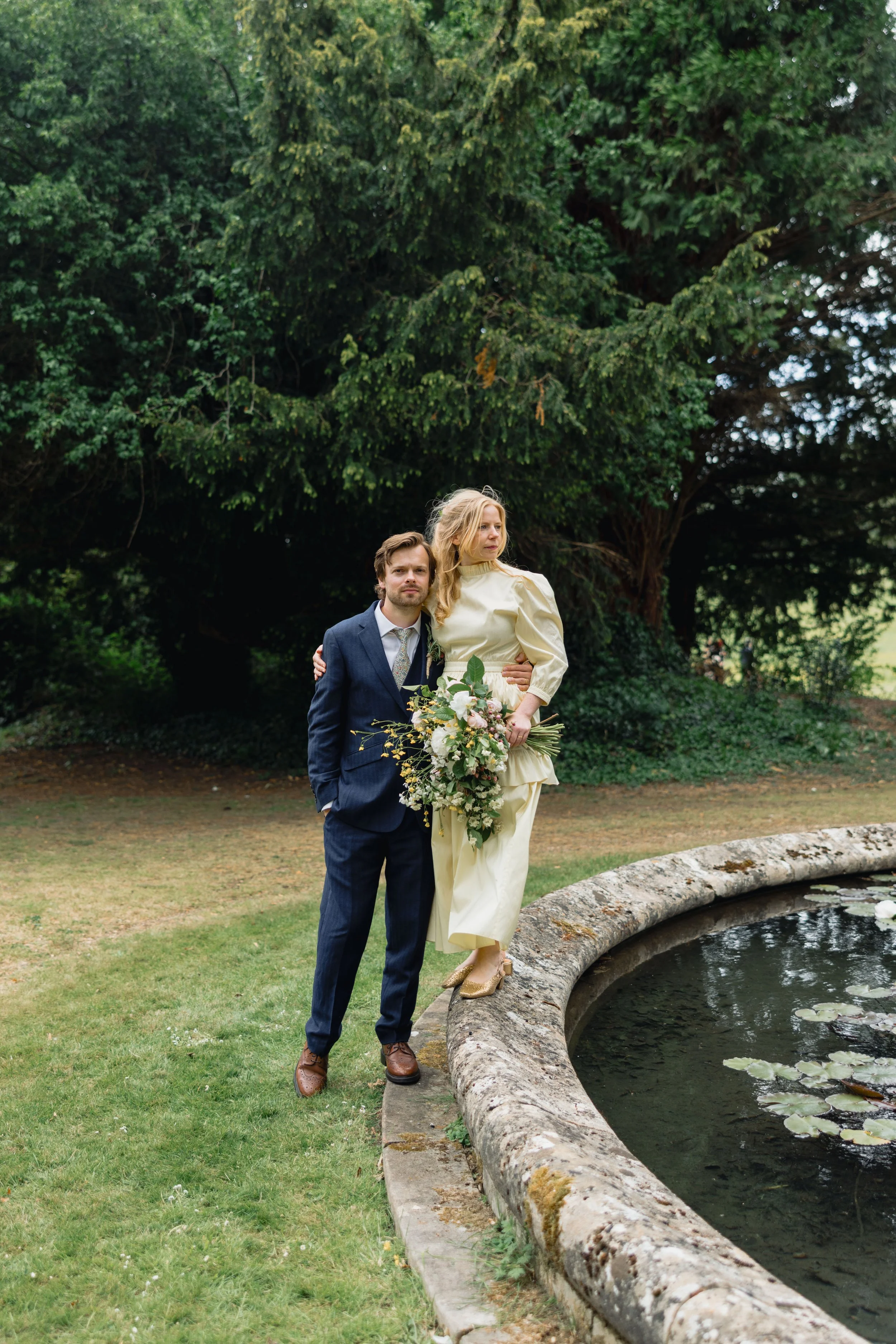 A bride and groom stand by a pond in a garden, with lush green trees in the background. The bride holds a bouquet of flowers and is wearing a cream-colored dress, while the groom wears a dark blue suit and brown shoes.
