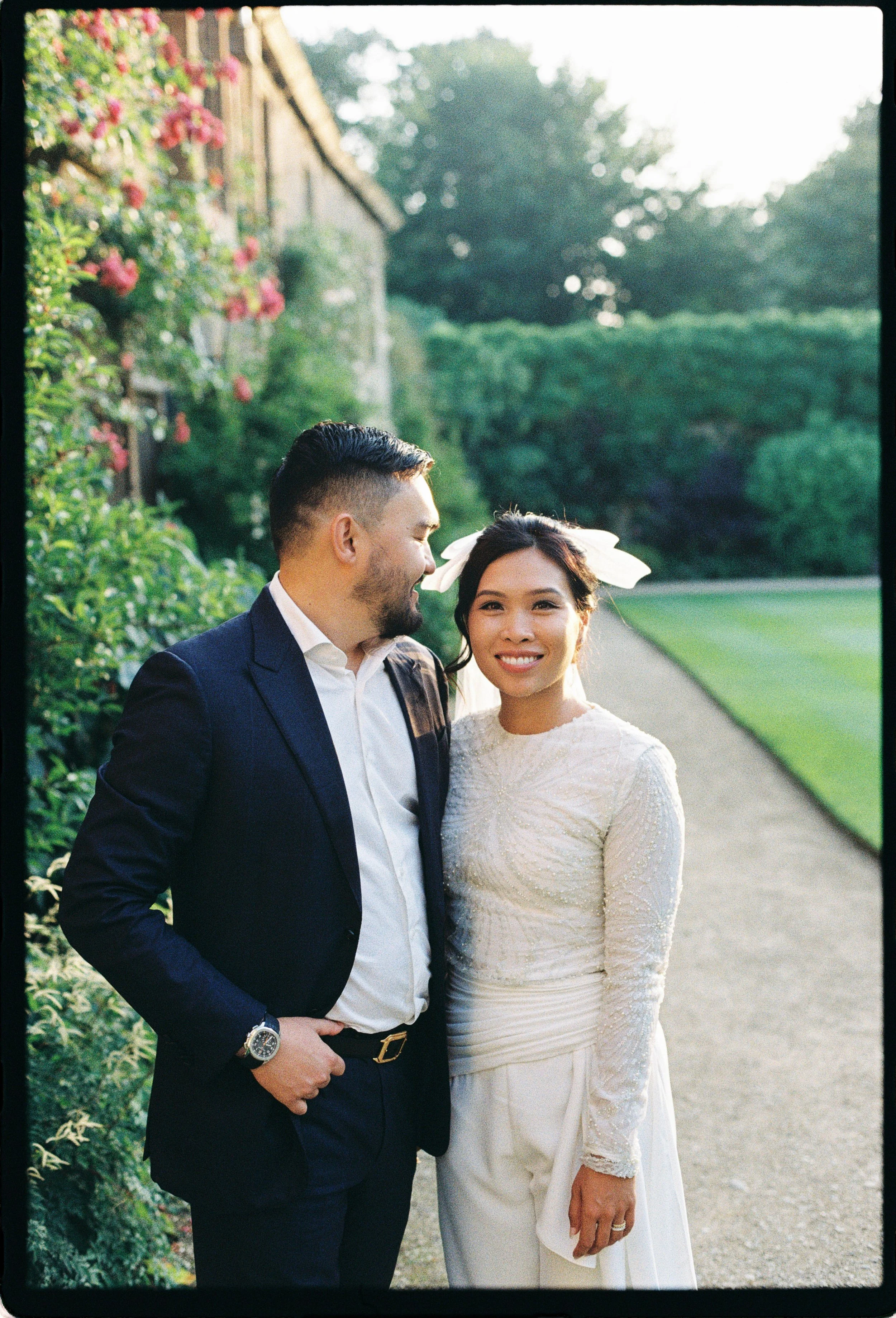 A smiling couple, a man in a dark suit and a woman in a white dress, standing outdoors on a garden pathway with greenery and pink flowers in the background.