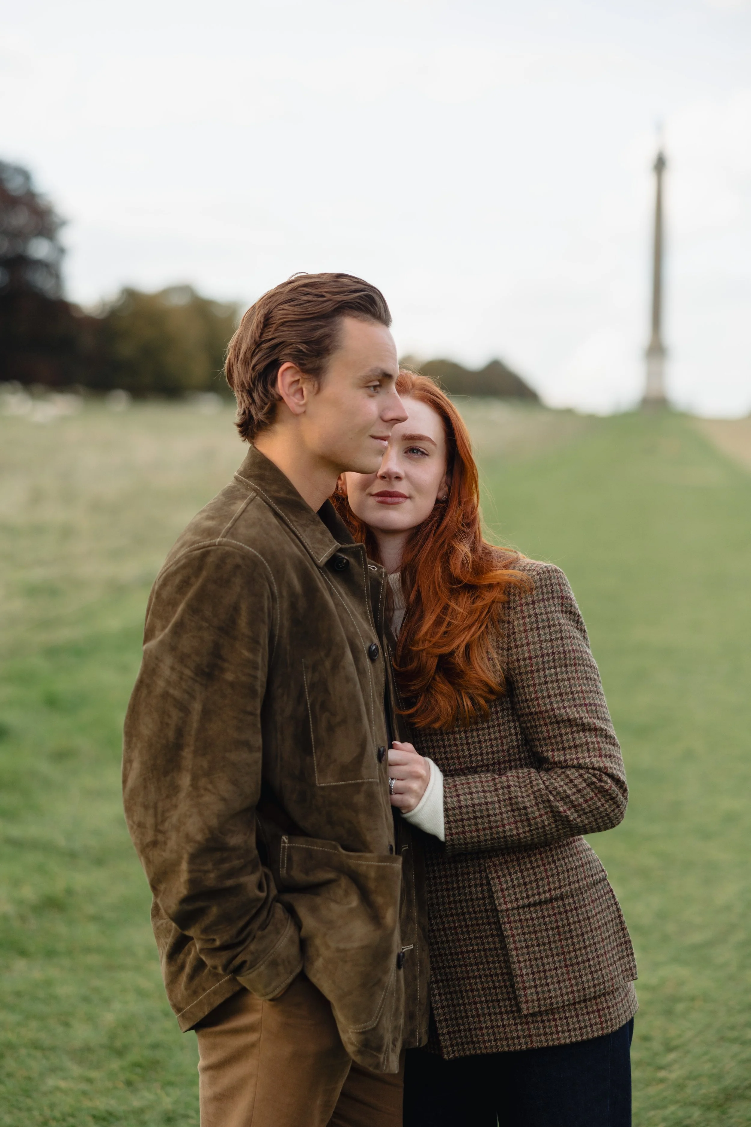 A young man and woman stand close together outdoors on a cloudy day, with a statue in the background, suggesting a park or historic site.