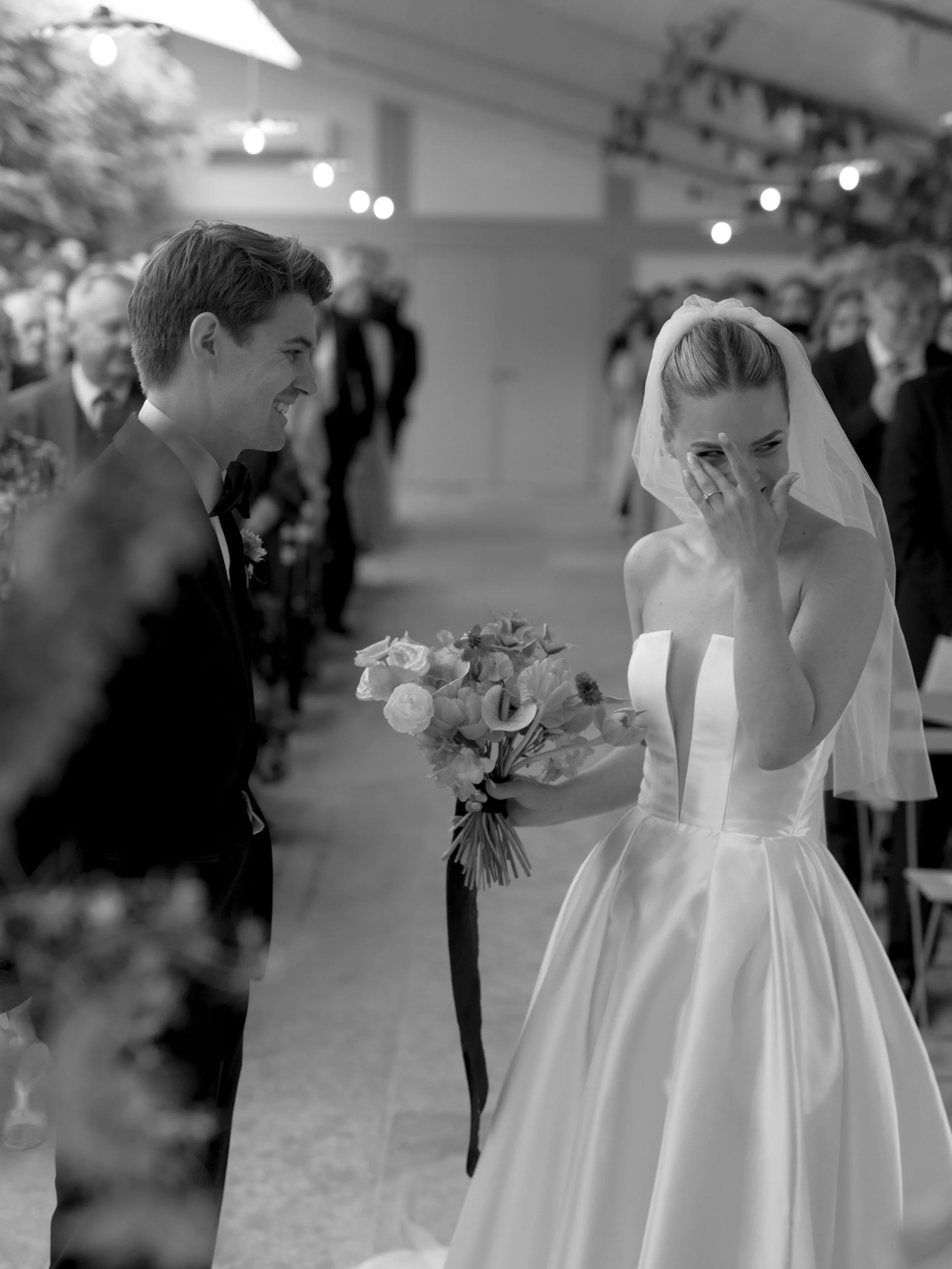 A bride in a strapless wedding gown holding a bouquet, covering her face and laughing, standing next to a groom in a tuxedo during a wedding ceremony at Wiltshire wedding venue, Kin House.