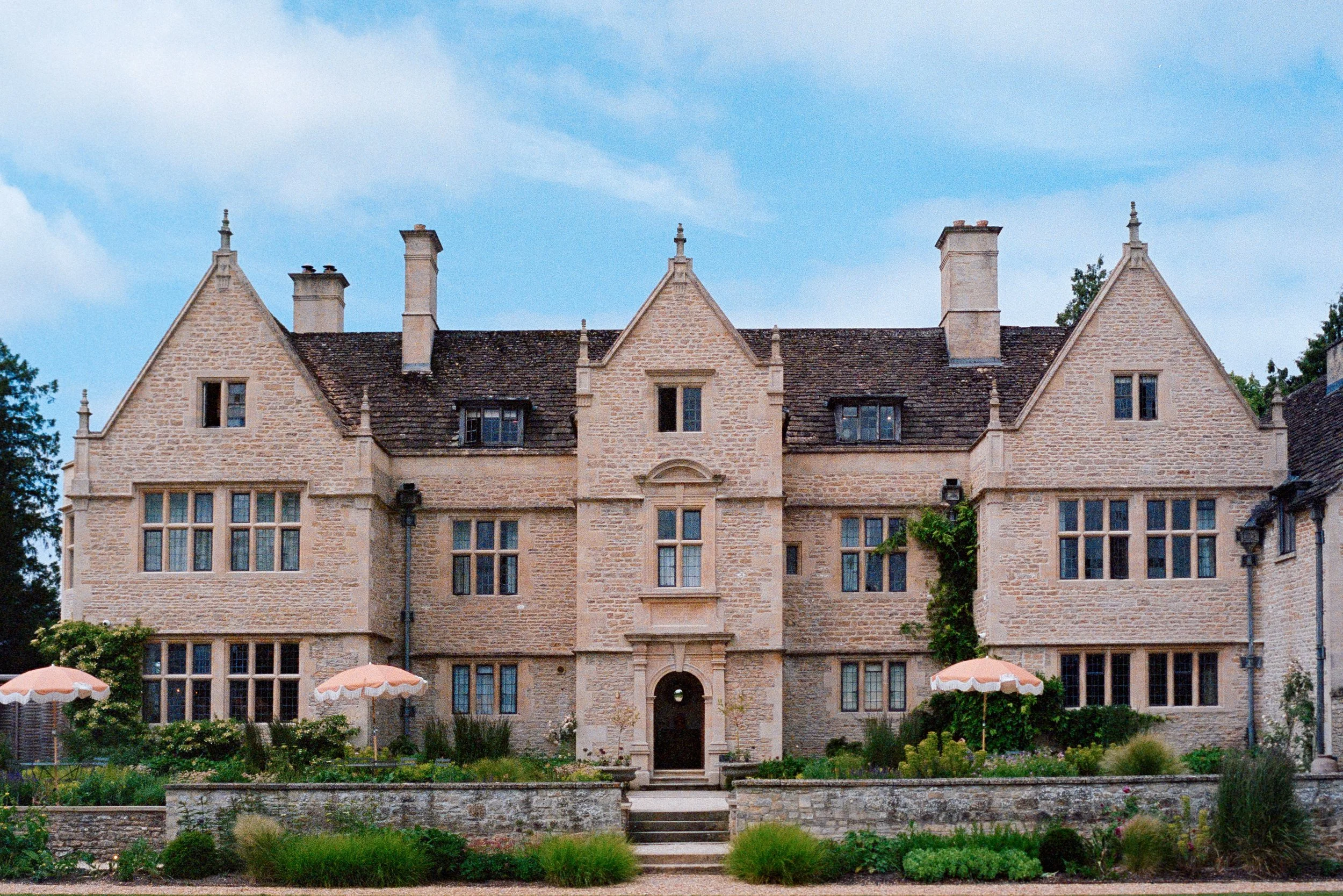 A large, historic stone mansion with multiple gabled roofs, tall chimneys, and numerous windows, surrounded by a well-maintained garden with umbrellas and outdoor seating.