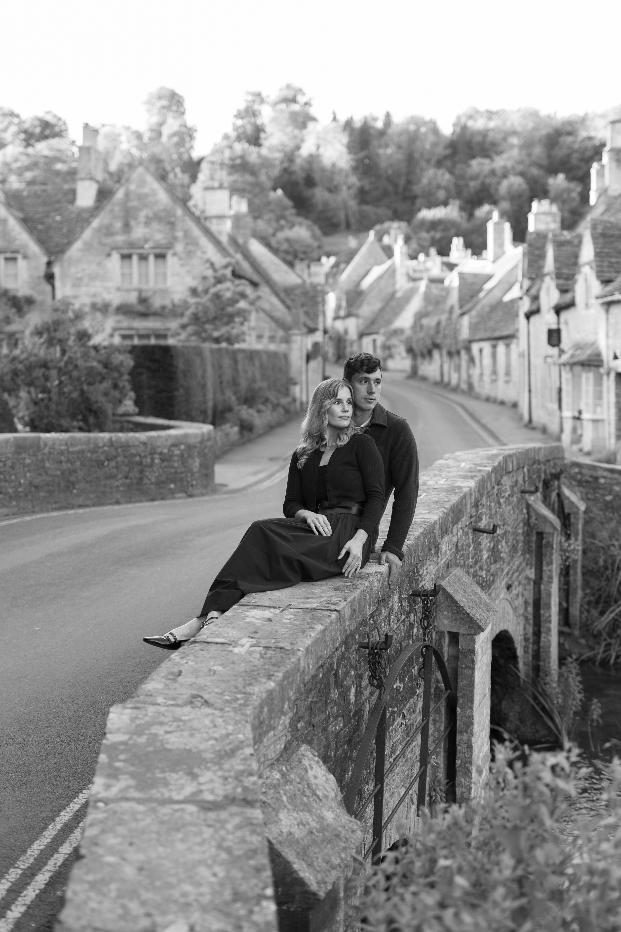 Black and white photo of a young woman and man sitting on a stone bridge, with a village street and houses in the background.