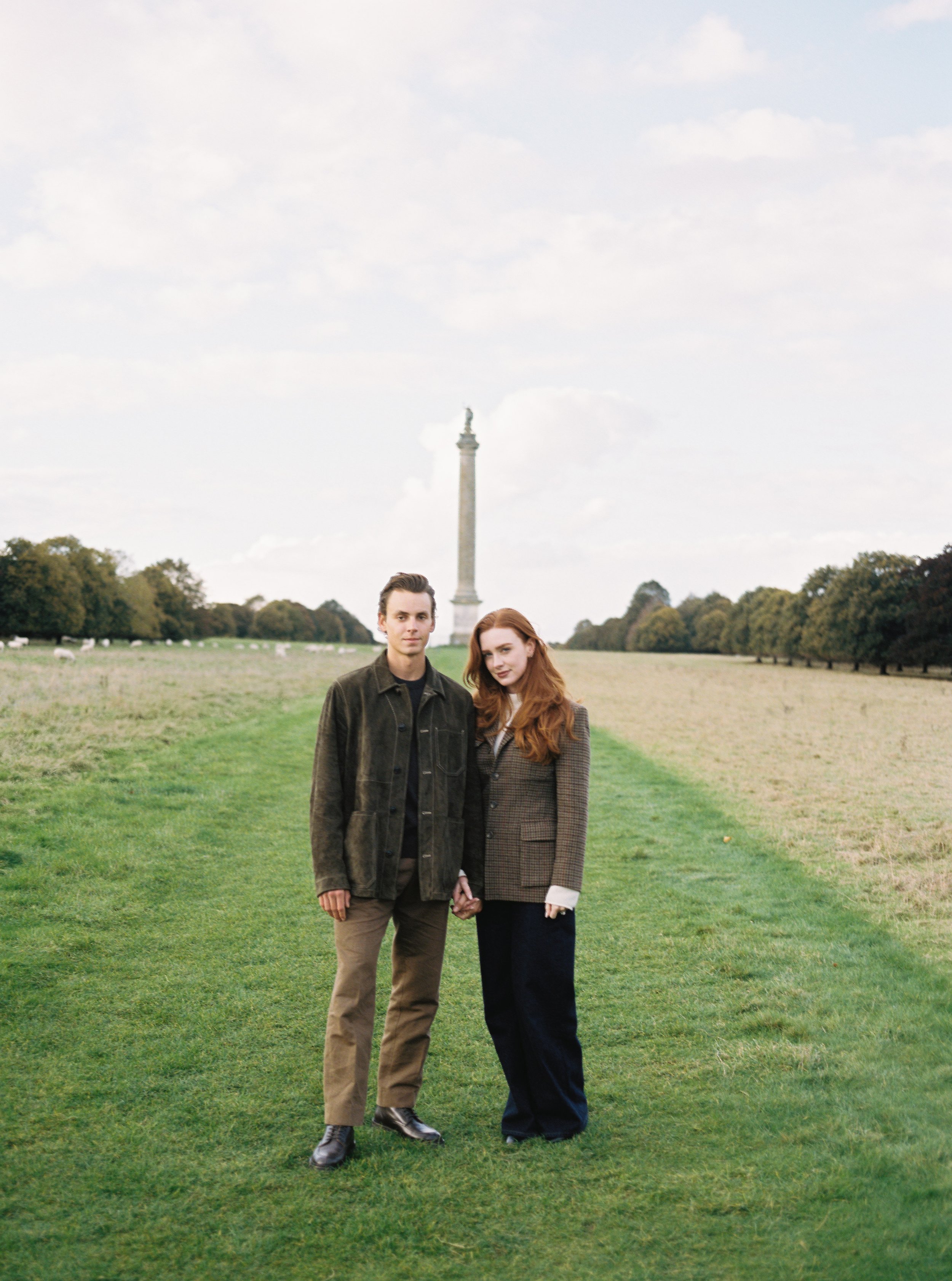A young couple holding hands stands on a grassy path with trees on both sides and a tall monument in the background.