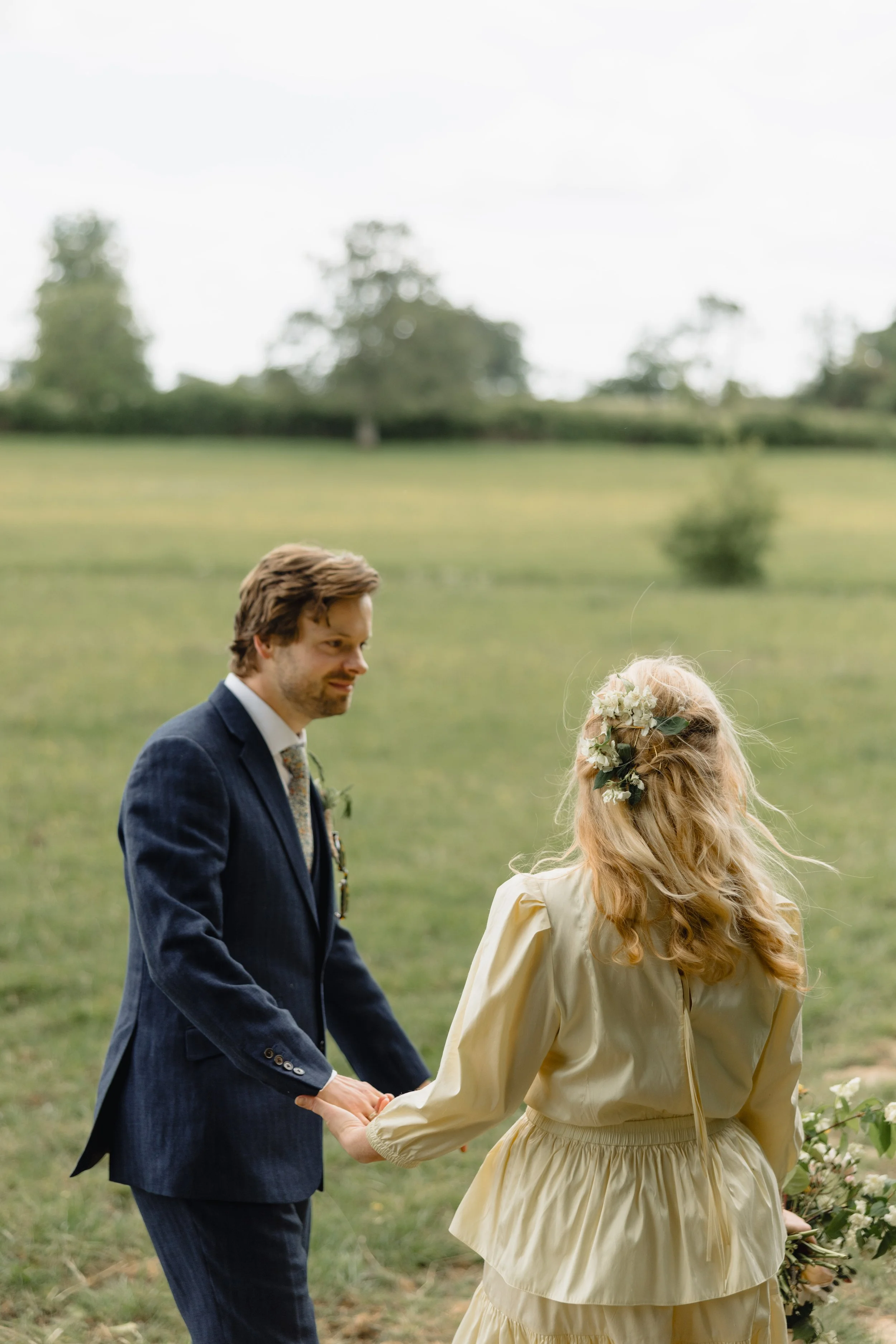 A groom and bride holding hands outdoors in a grassy field during a wedding ceremony, with trees and an overcast sky in the background.