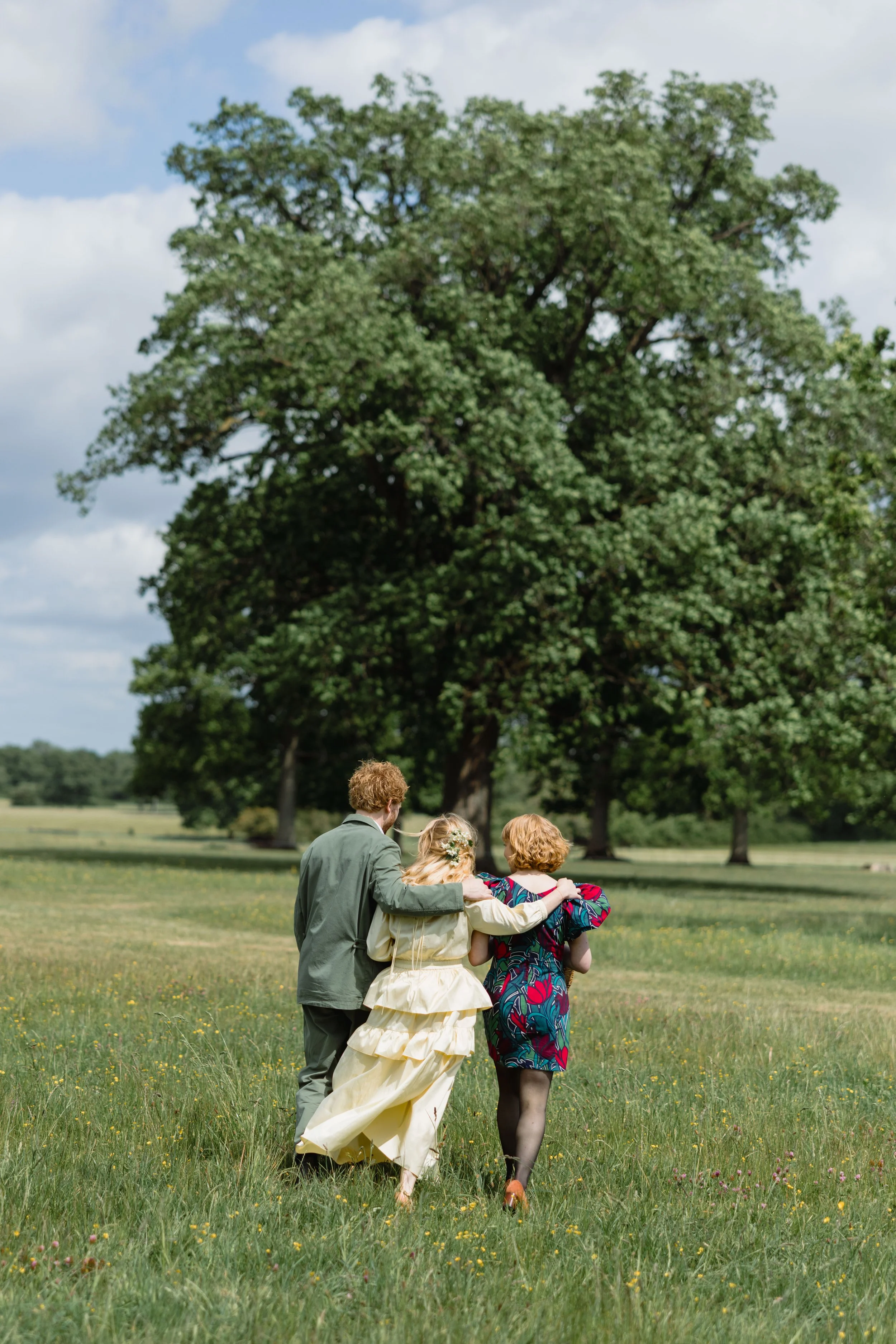 Three people walking on grass in a park with large trees and partly cloudy sky.