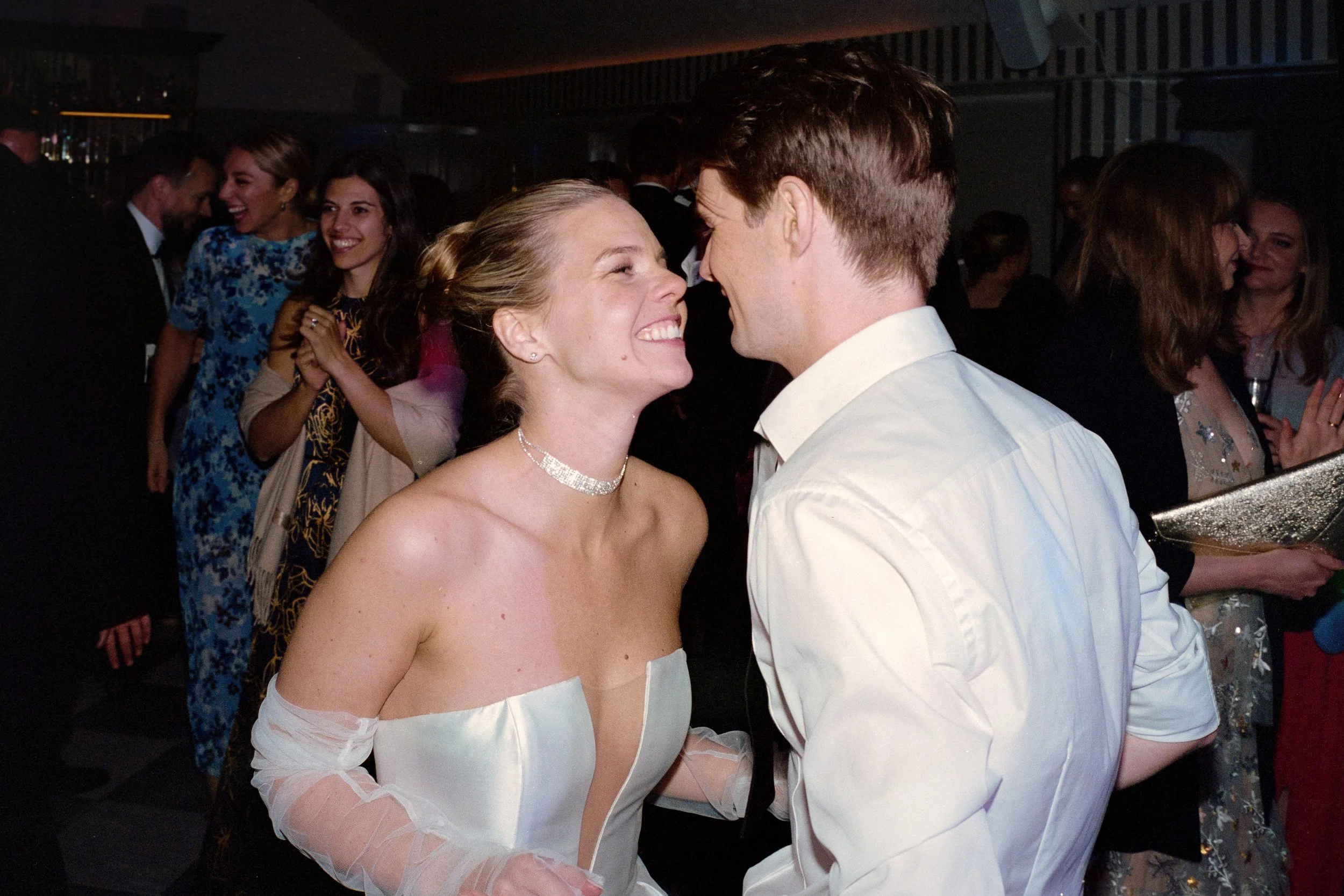 A bride and groom smiling and leaning close together during their wedding reception, surrounded by guests socializing and celebrating.