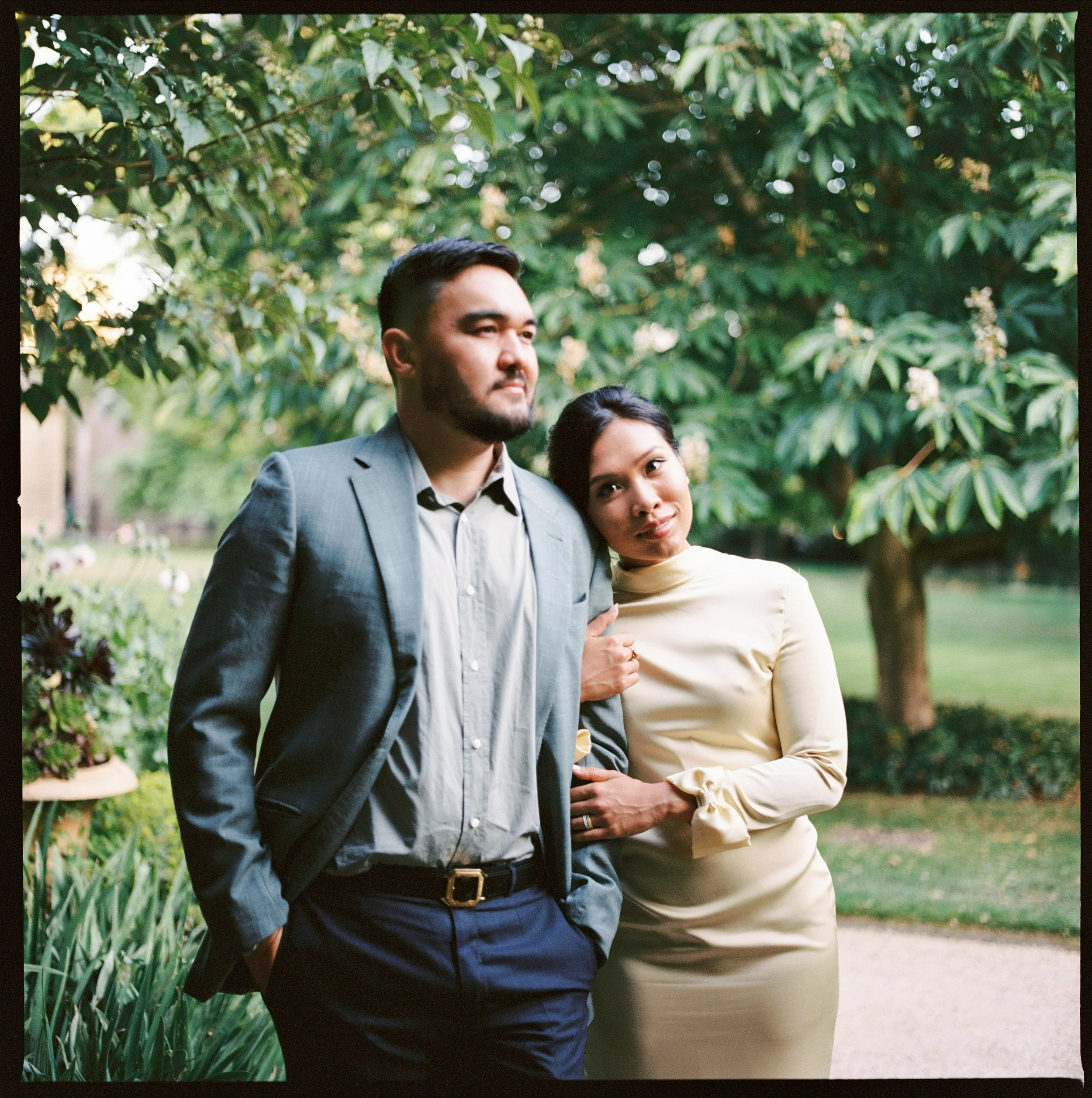 A man in a gray suit jacket and a woman in a cream-colored dress standing close together outdoors with green trees and plants in the background