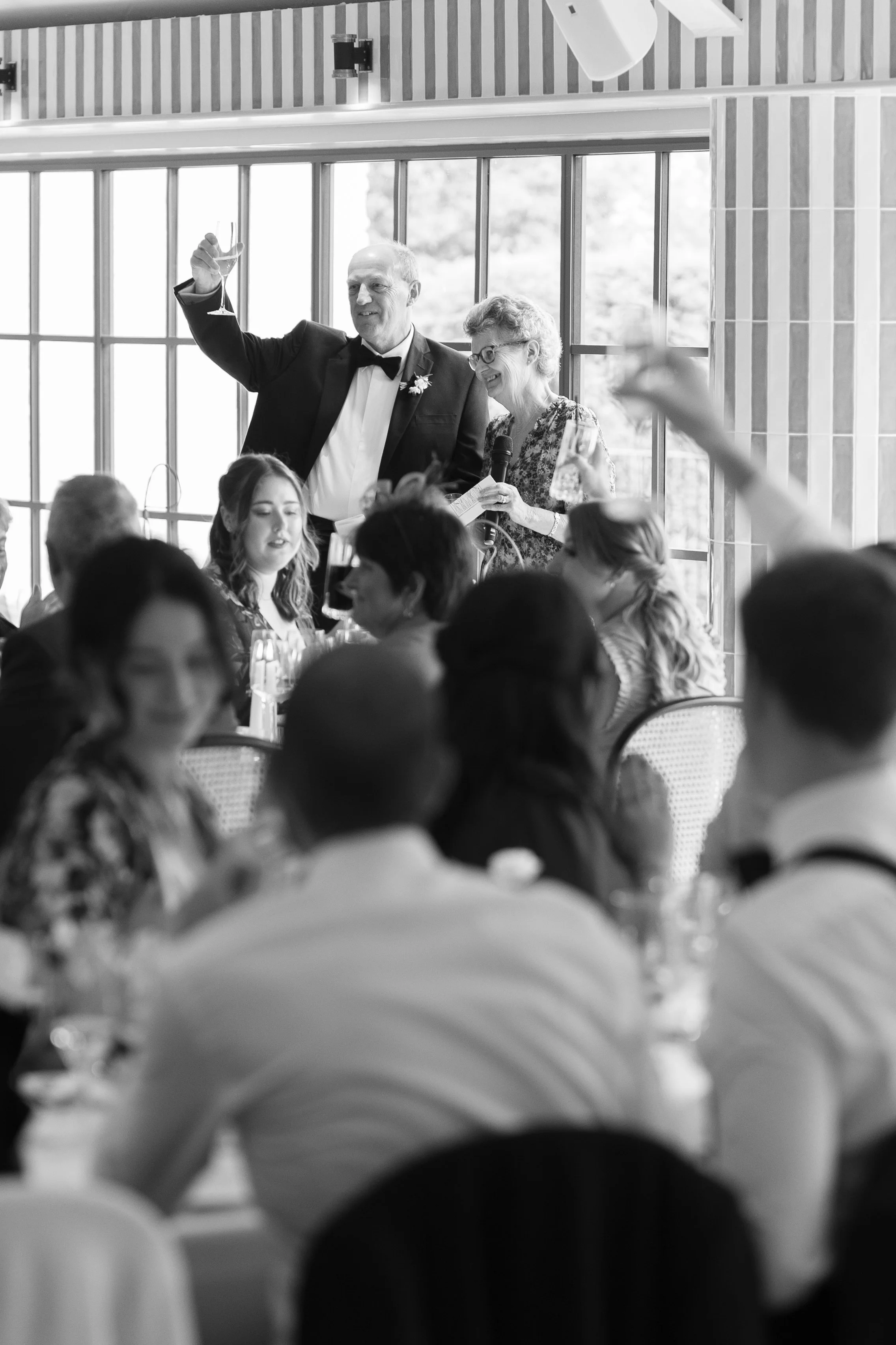 An older man in a tuxedo and an older woman with glasses are giving a toast at a wedding reception. The man is raising a glass, and the woman is holding a microphone and a glass. Guests seated at tables are watching and smiling.